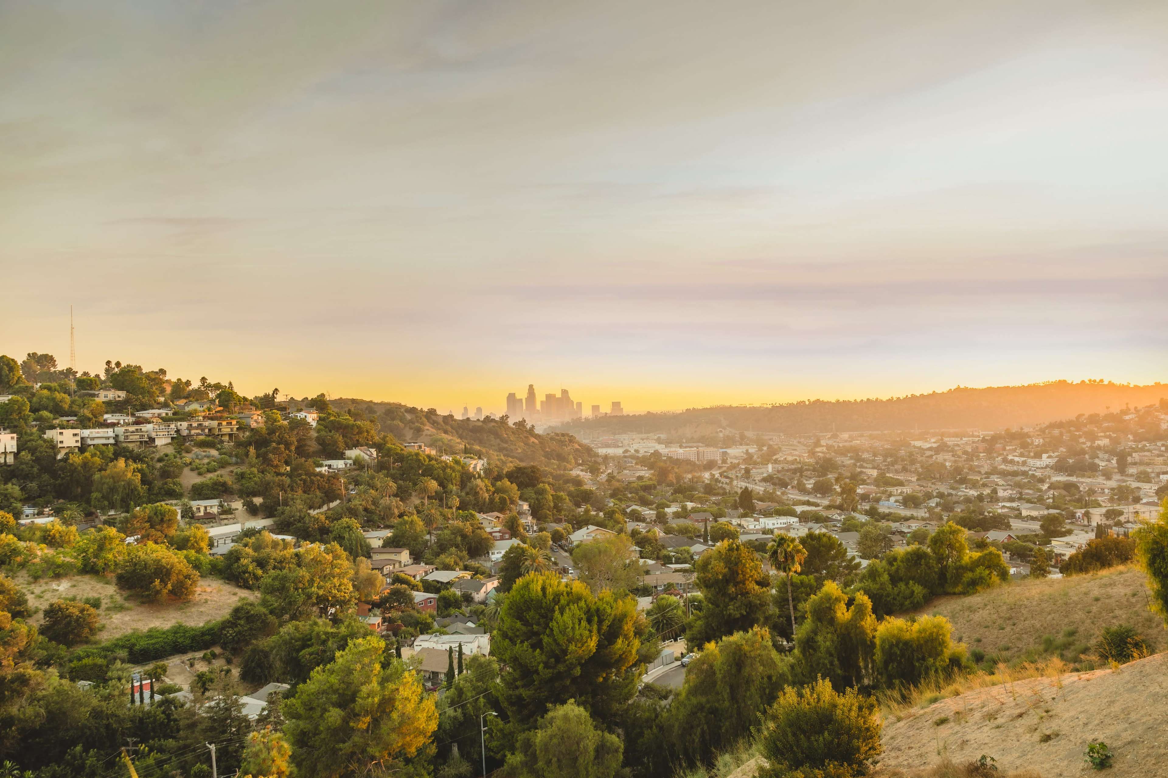 The image shows a sunset view of a hilly Los Angeles landscape, with the city's skyline visible in the background.
