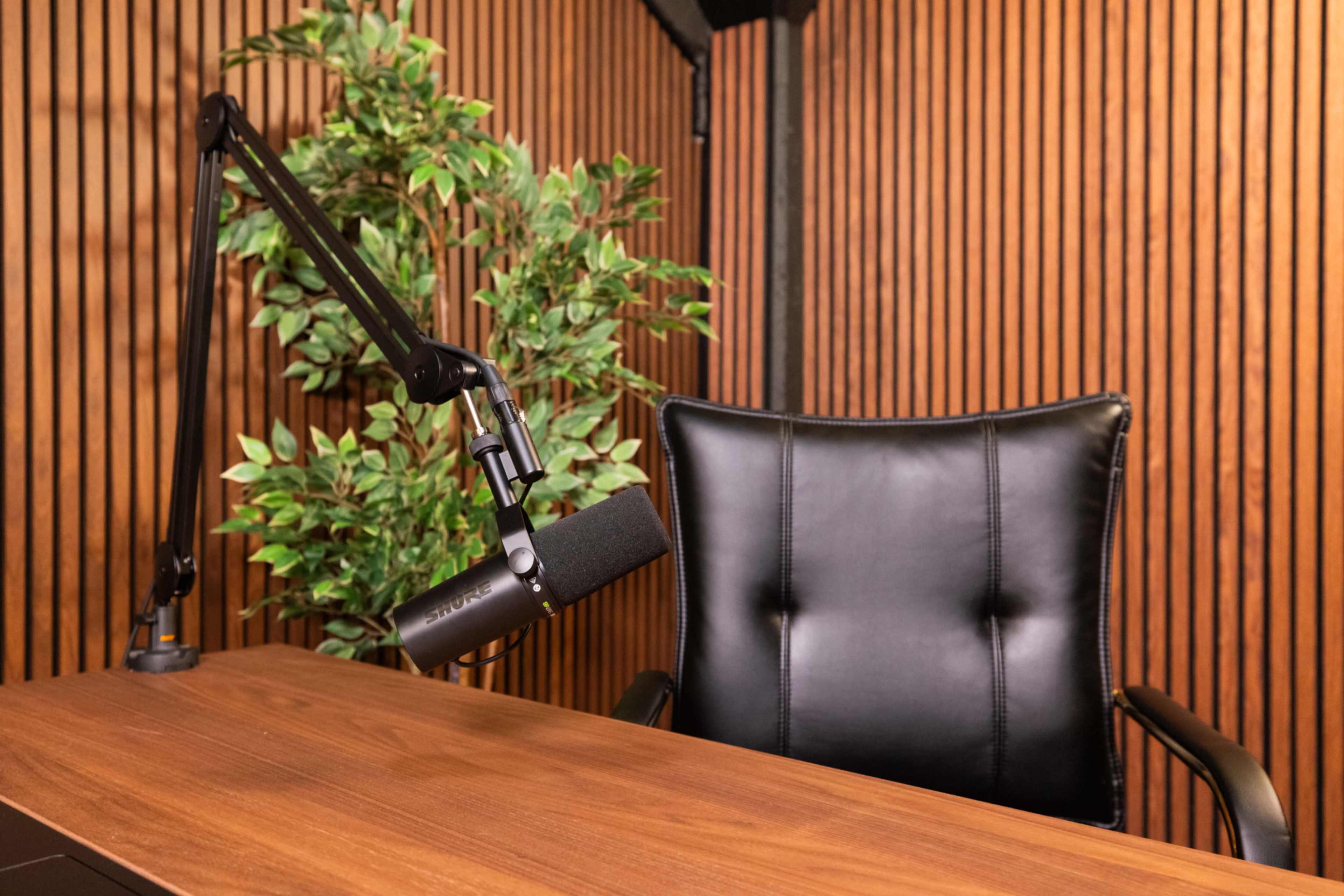 A black office chair with a microphone attached to a boom arm is positioned next to a wooden desk, set against a backdrop of wooden paneling and a potted plant.