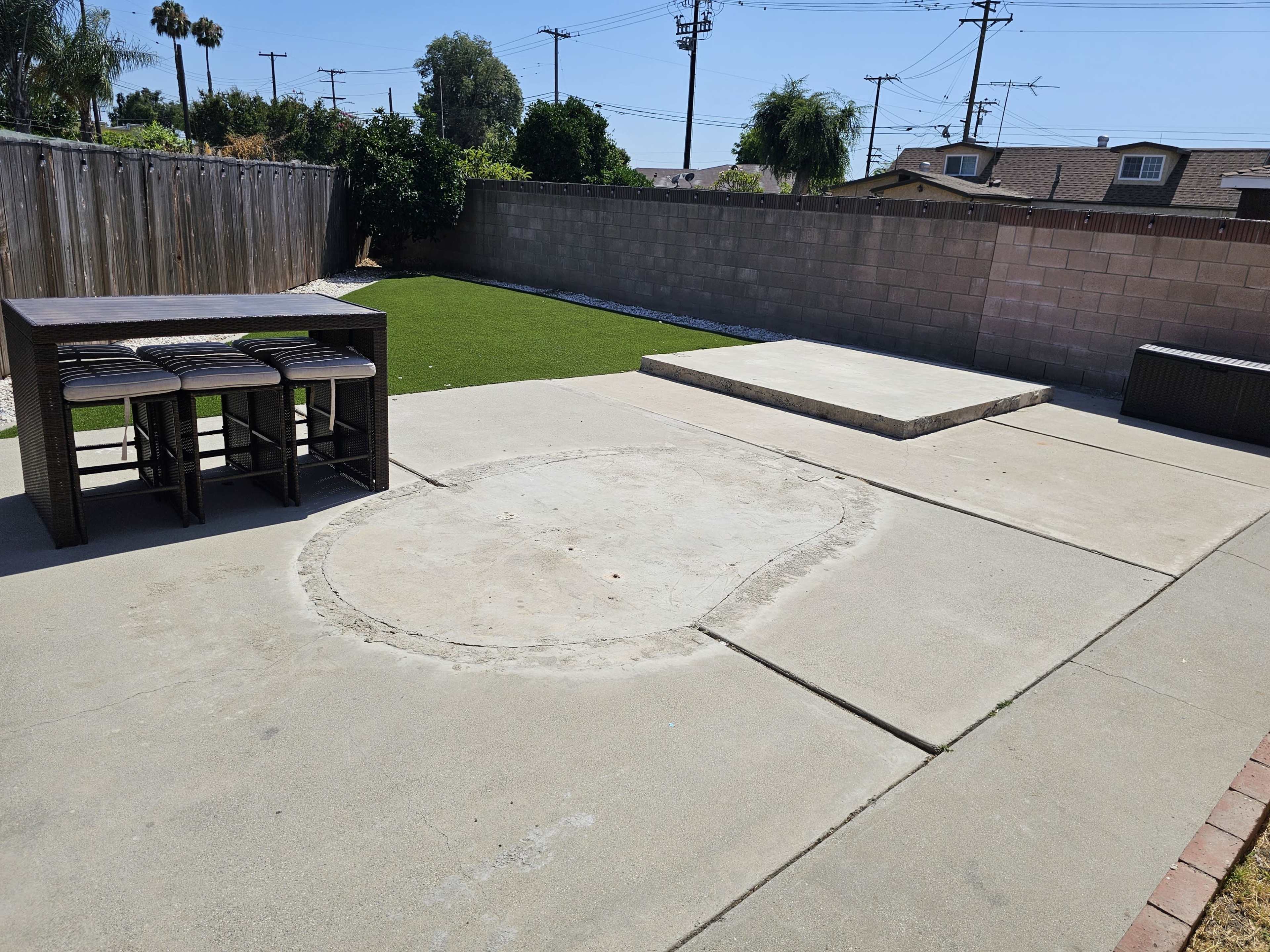 A backyard with a concrete patio, a set of four bar stools, a circular area that appears to be incomplete, and a raised platform at the far end, surrounded by a low wall and green grass.