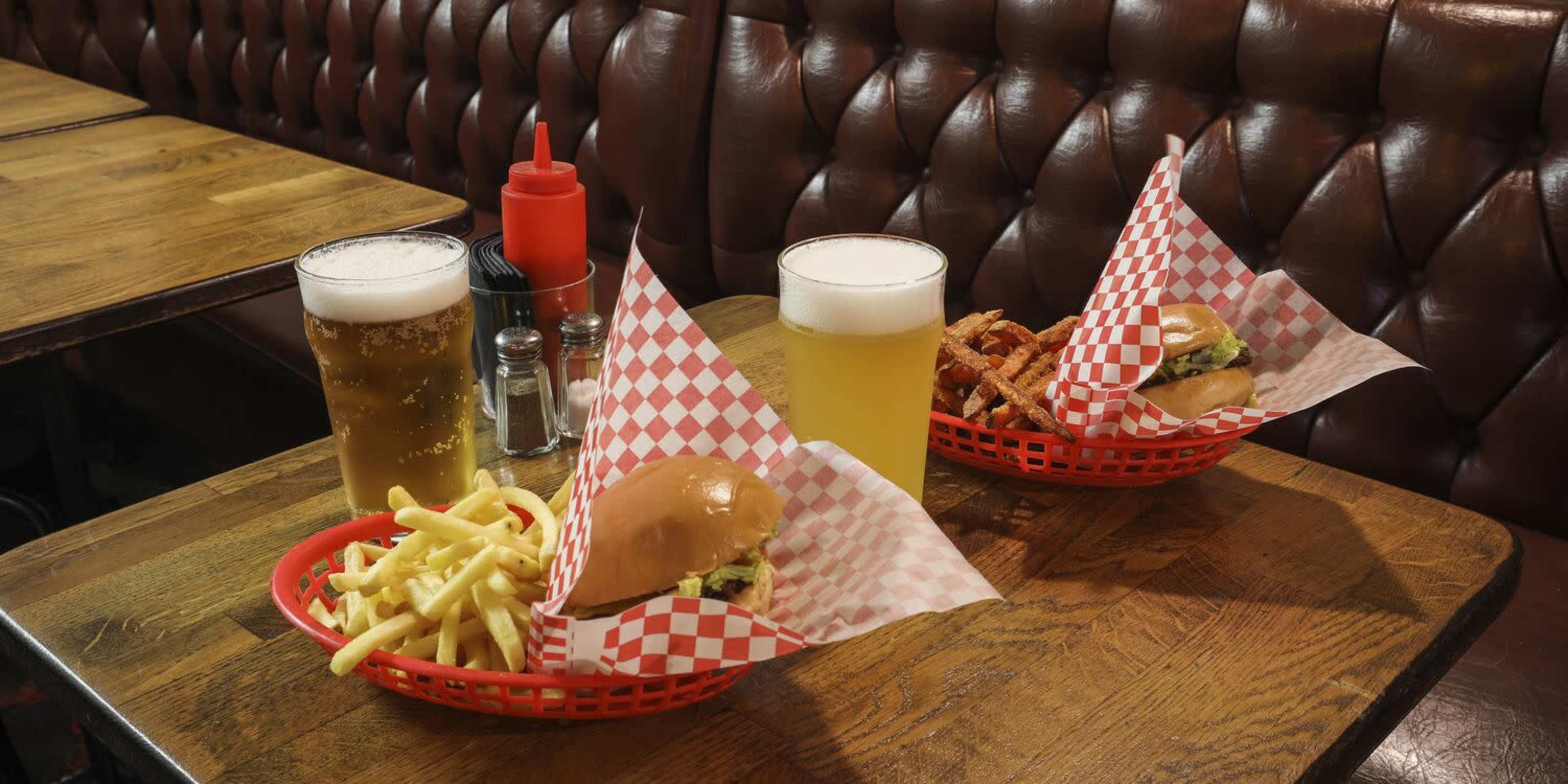Two baskets of burgers and fries, along with three glasses of beer, are placed on a wooden table in a cushioned booth setting.