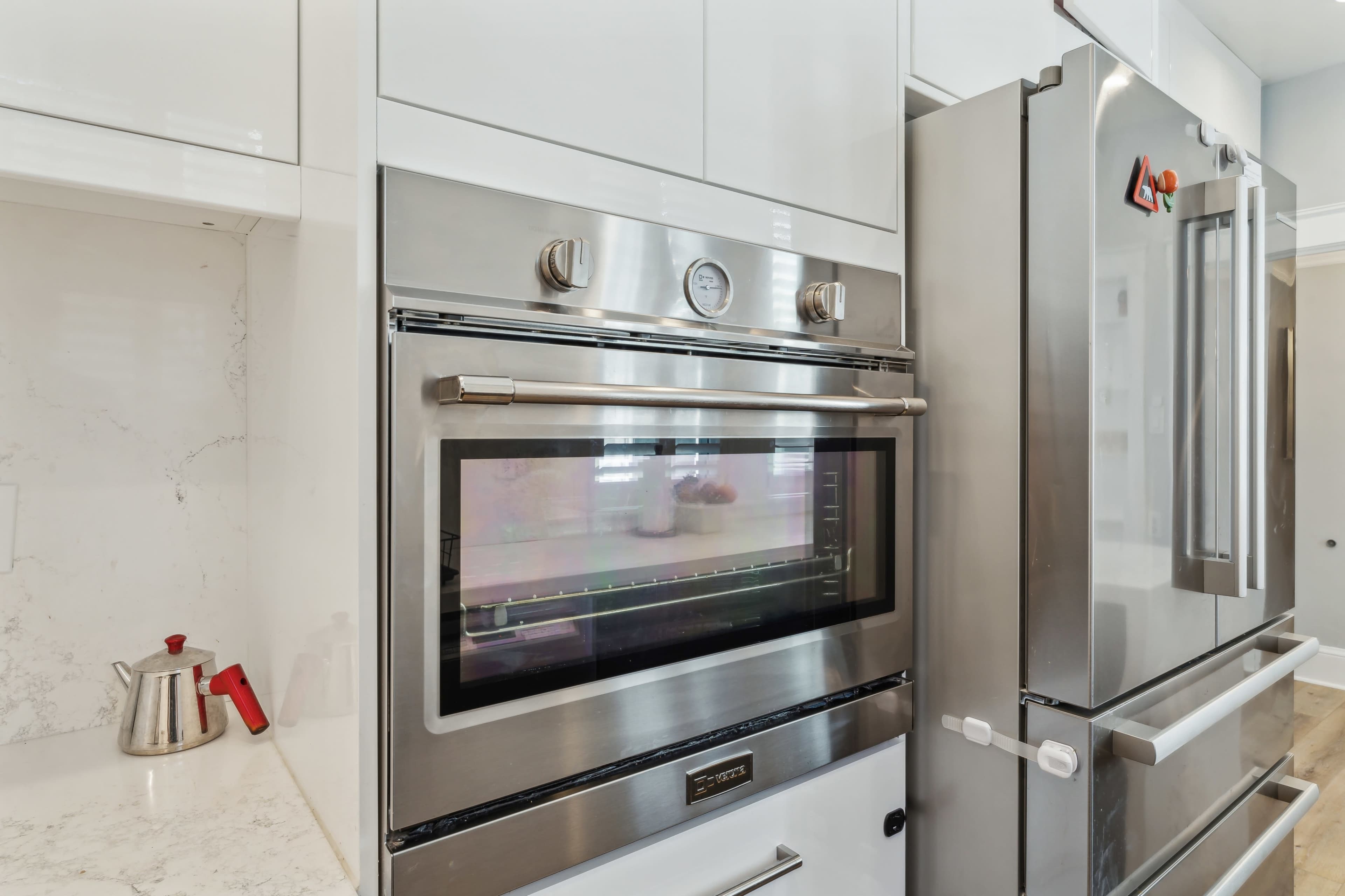 A modern kitchen with a stainless steel oven built into white cabinetry next to a silver refrigerator.