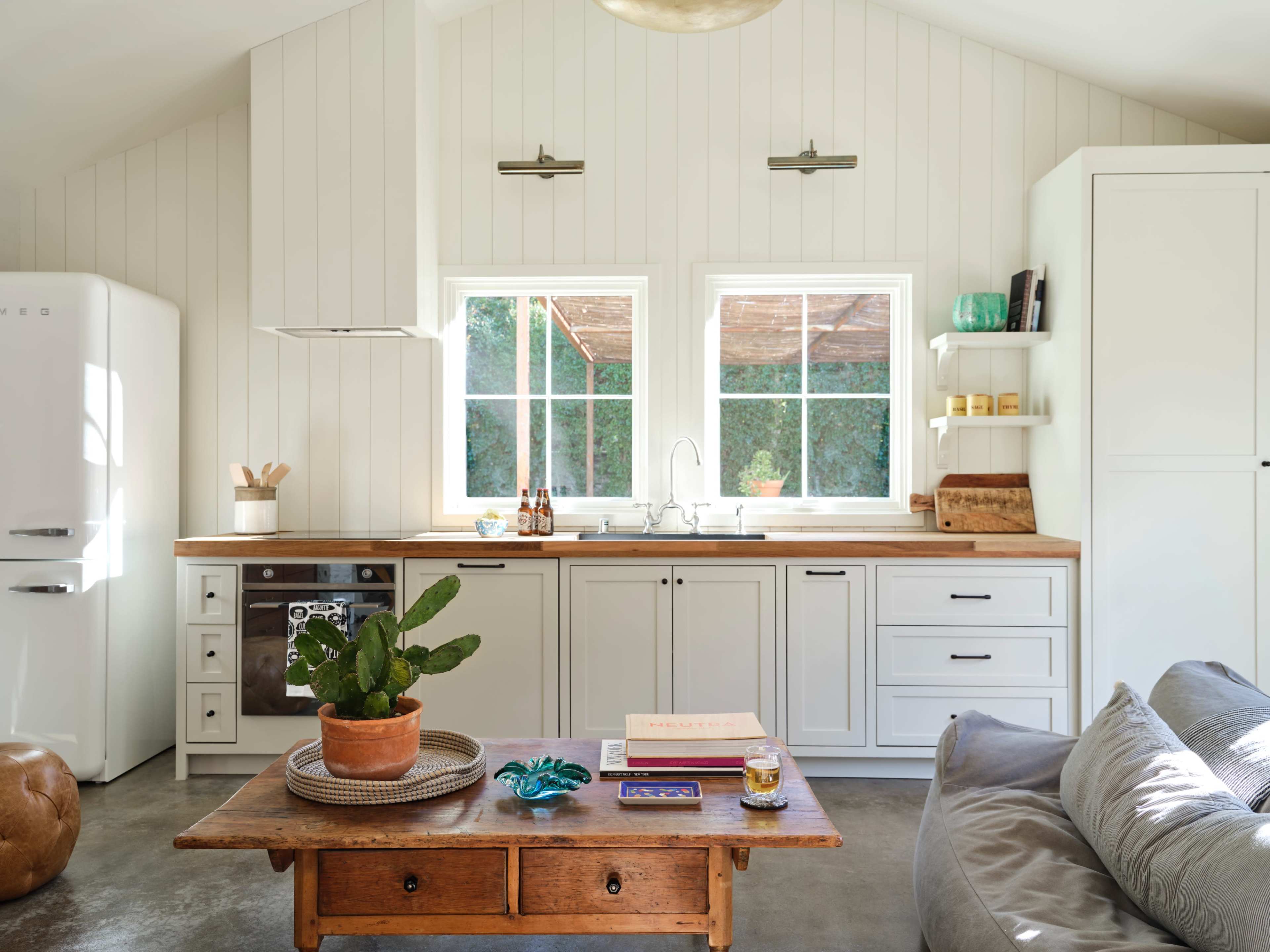 The kitchen features white shiplap walls, large windows, wooden countertops, and a central wooden table with a potted plant.
