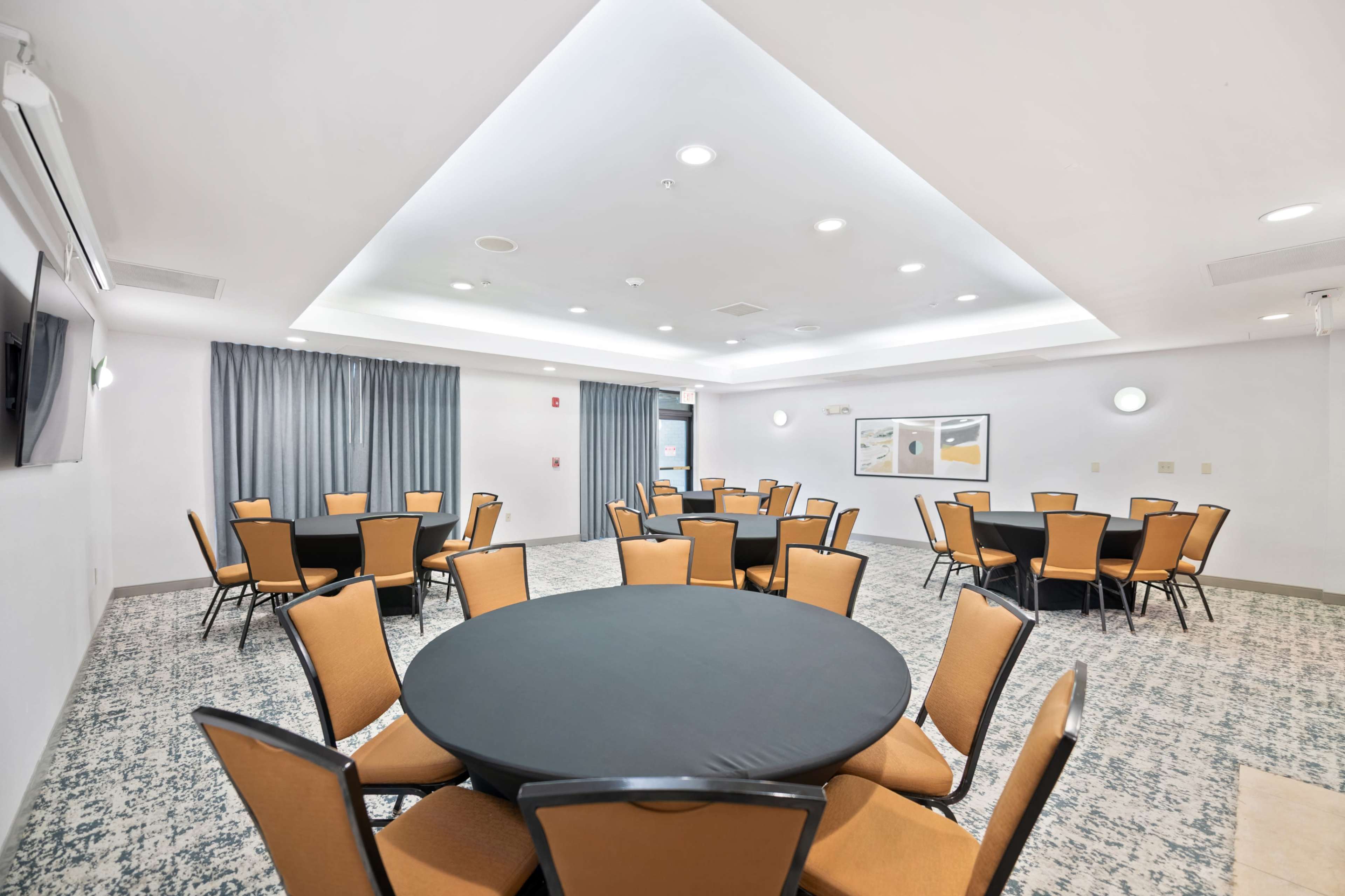 The image shows a well-lit meeting room featuring several round tables with black tablecloths and orange chairs arranged for an event.