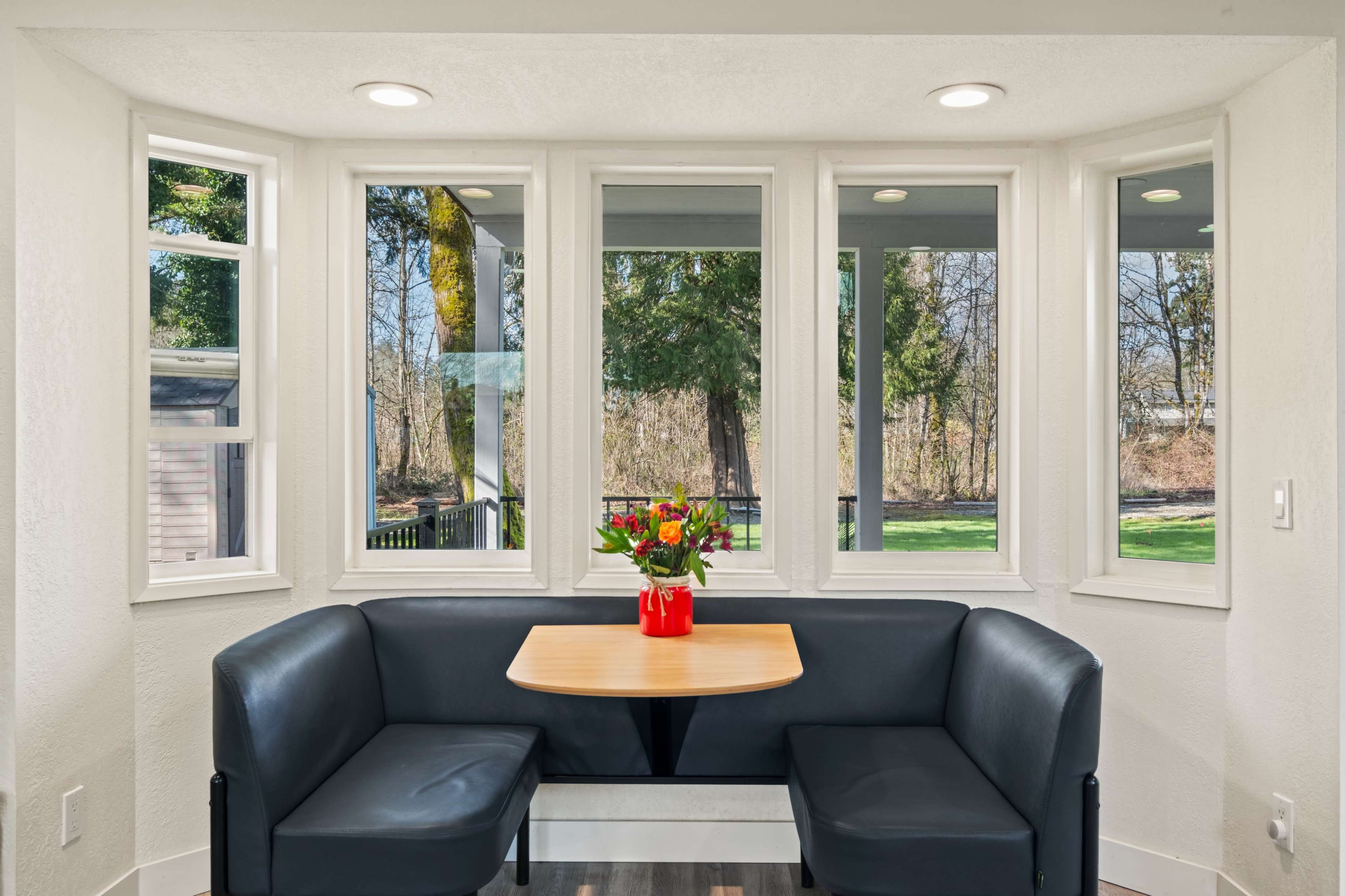 A cozy nook with a round table and a flower arrangement is surrounded by large windows offering a view of the outdoors.