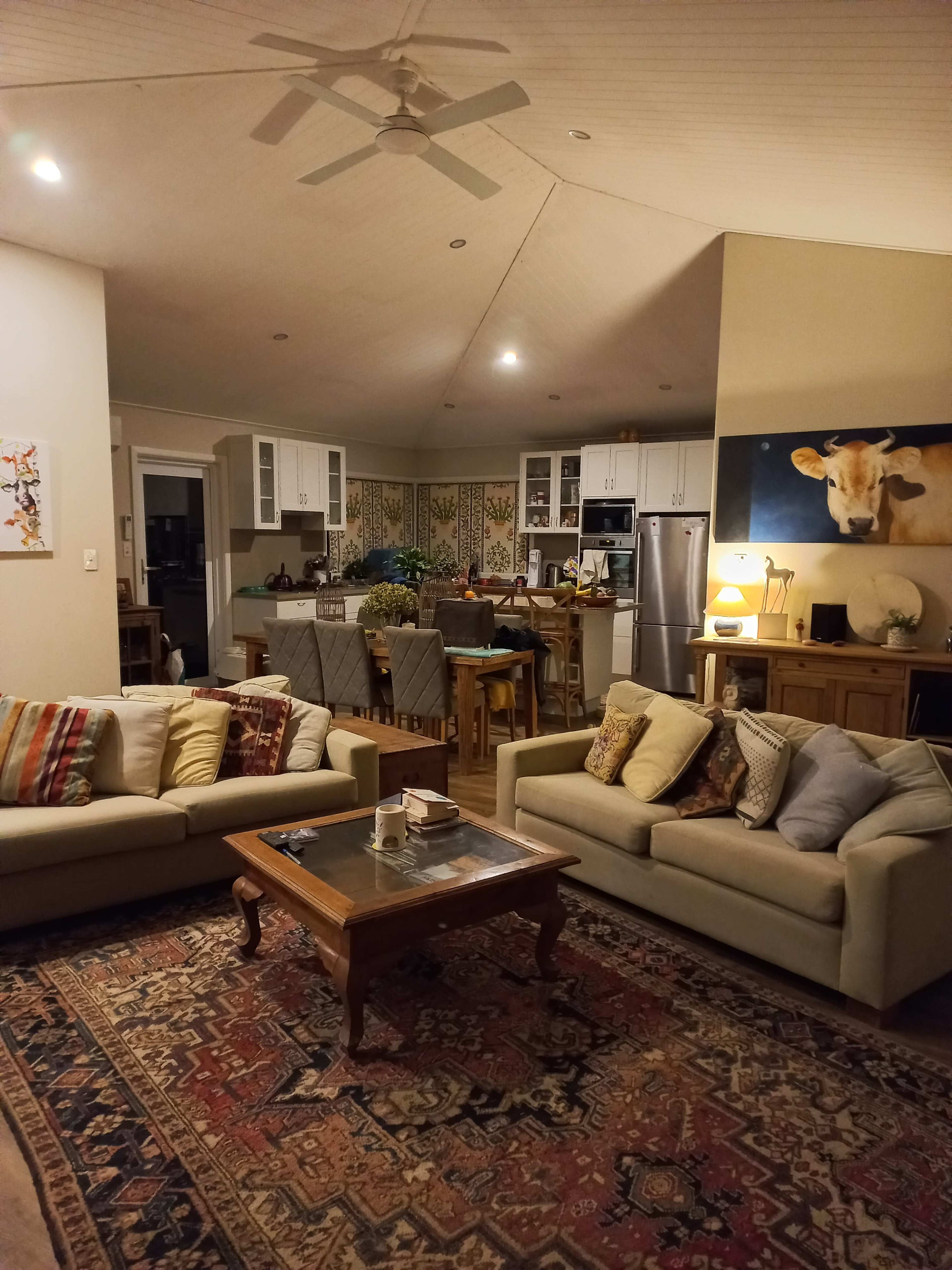 A cozy living room features two sofas, a wooden coffee table, and an open kitchen in the background with light-colored cabinetry and a large painting of a cow on the wall.