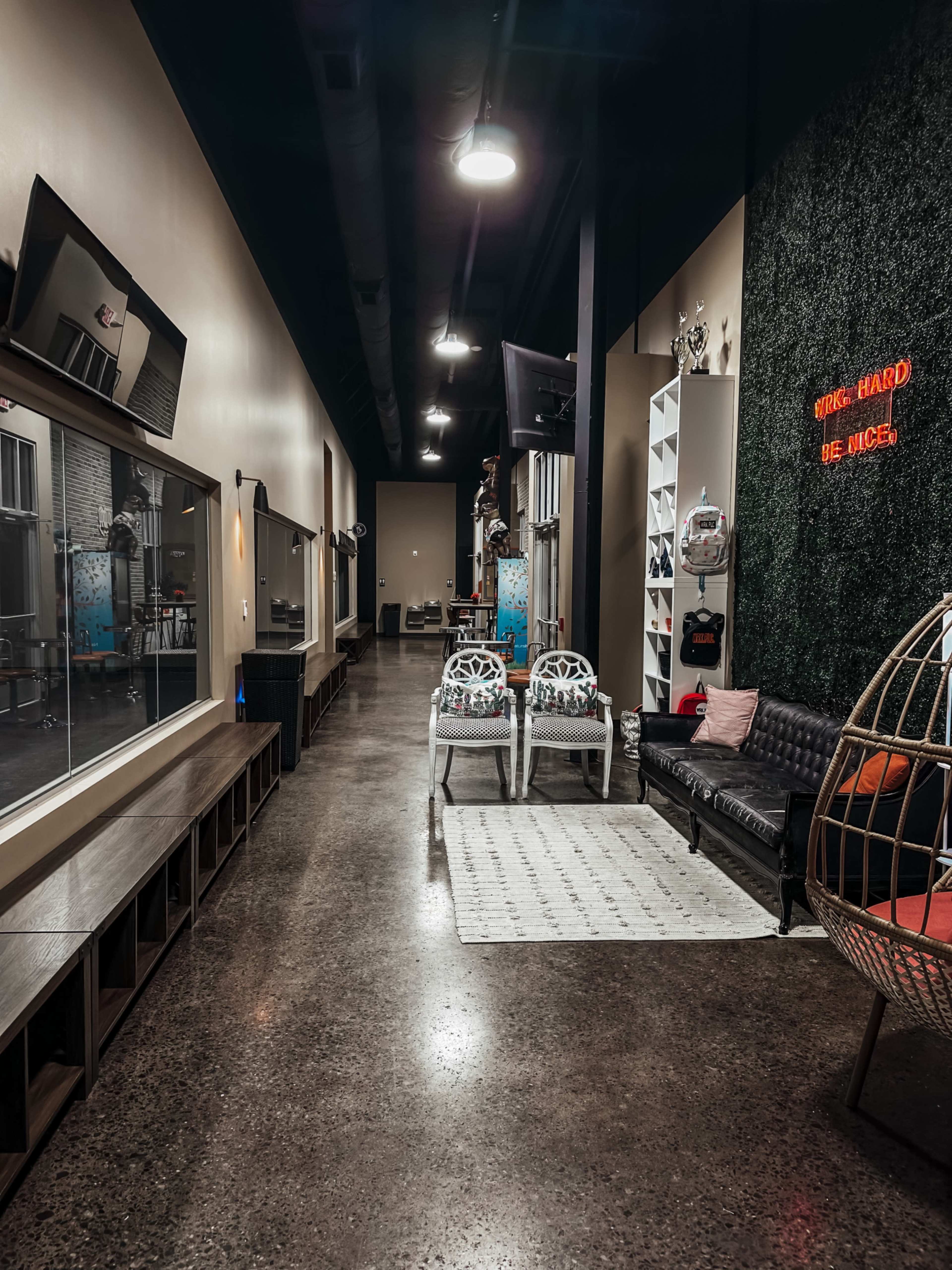 The image shows a modern hallway with polished concrete floors, lined with seating areas and decor, including a neon sign that reads "WORK HARD BE NICE."