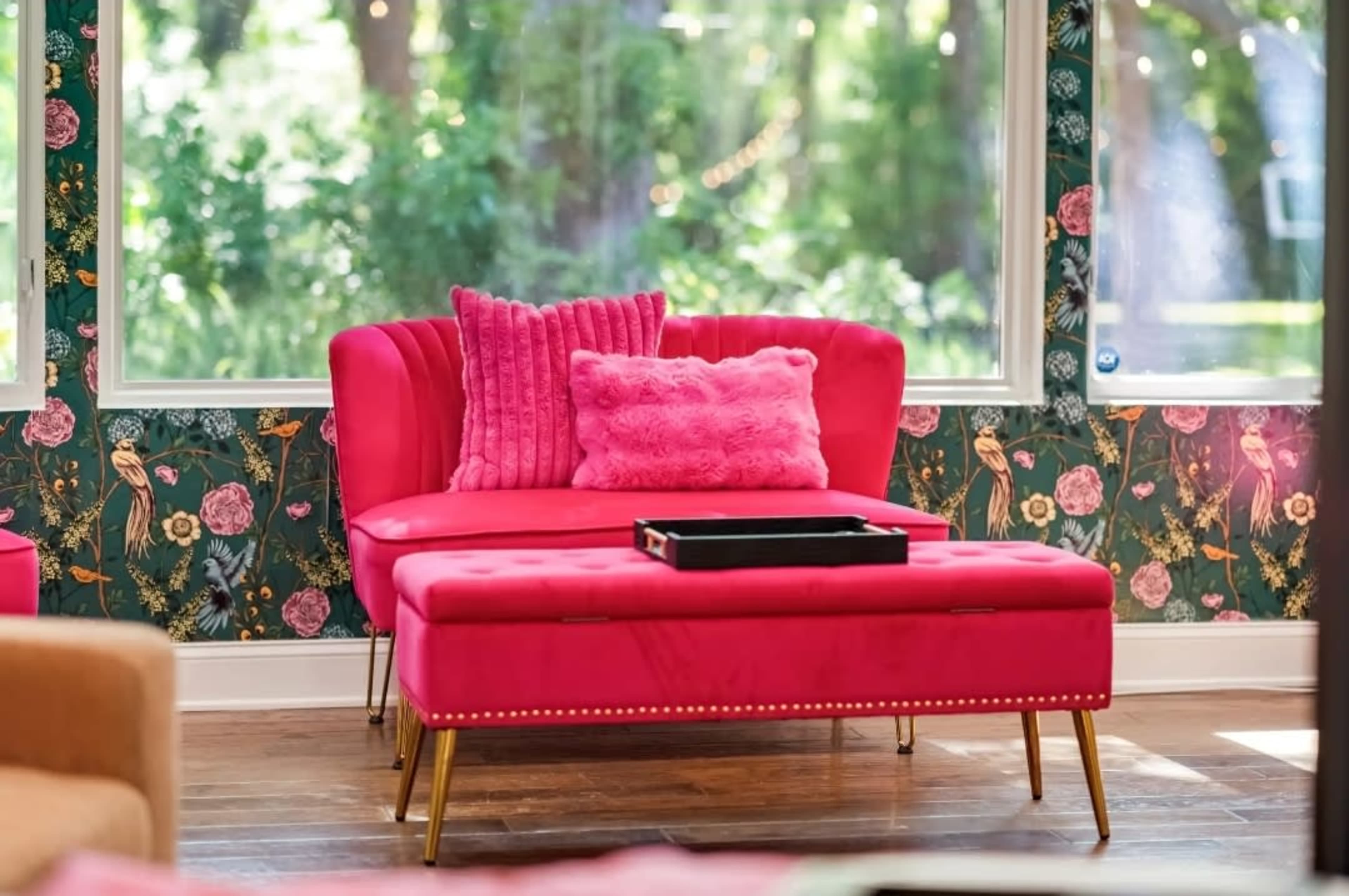 A bright pink couch and matching ottoman are set against a floral wallpaper background with large windows providing natural light.