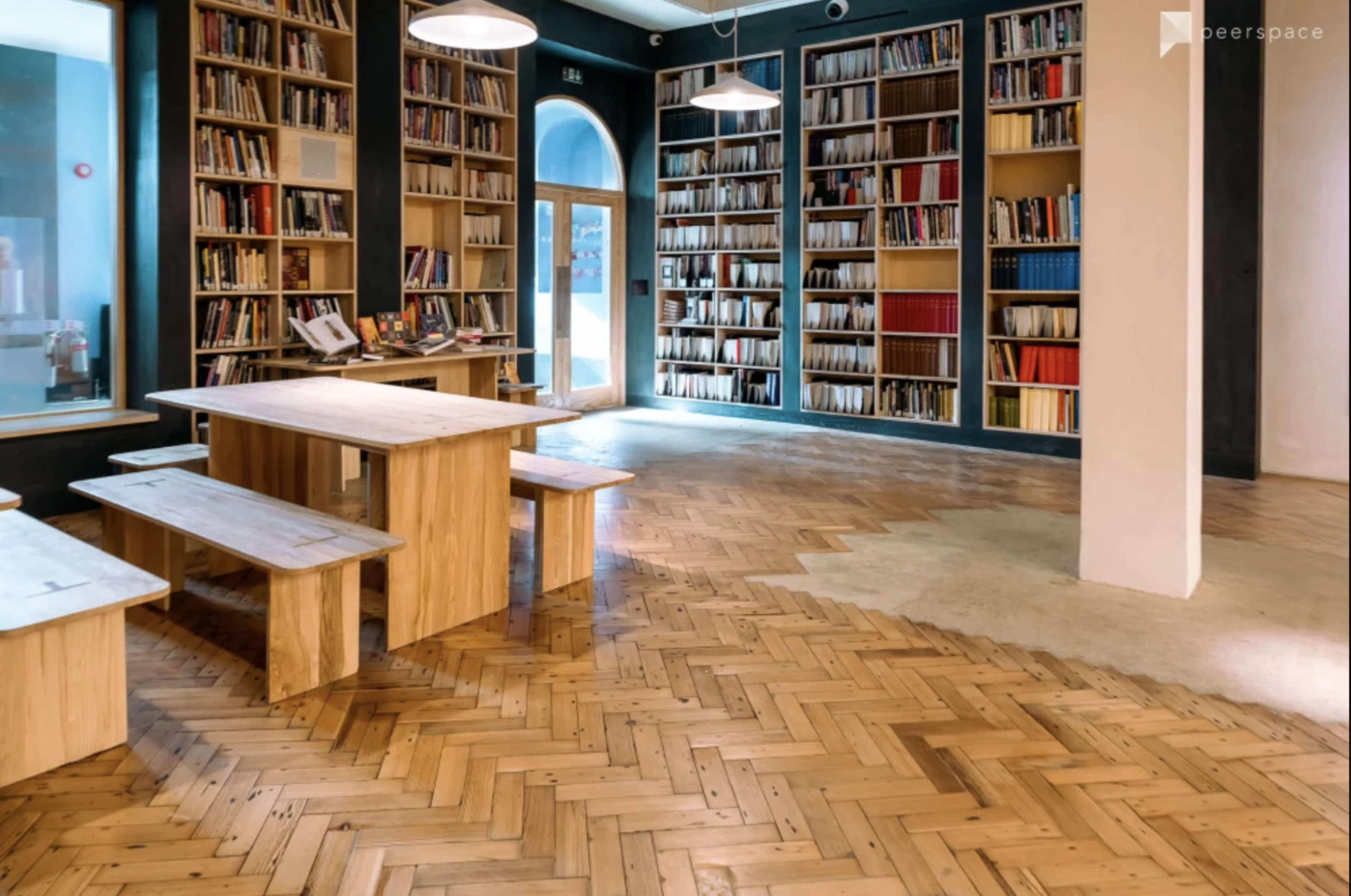 A modern library interior featuring tall shelves filled with books, a wooden table with benches, and a herringbone pattern wooden floor.