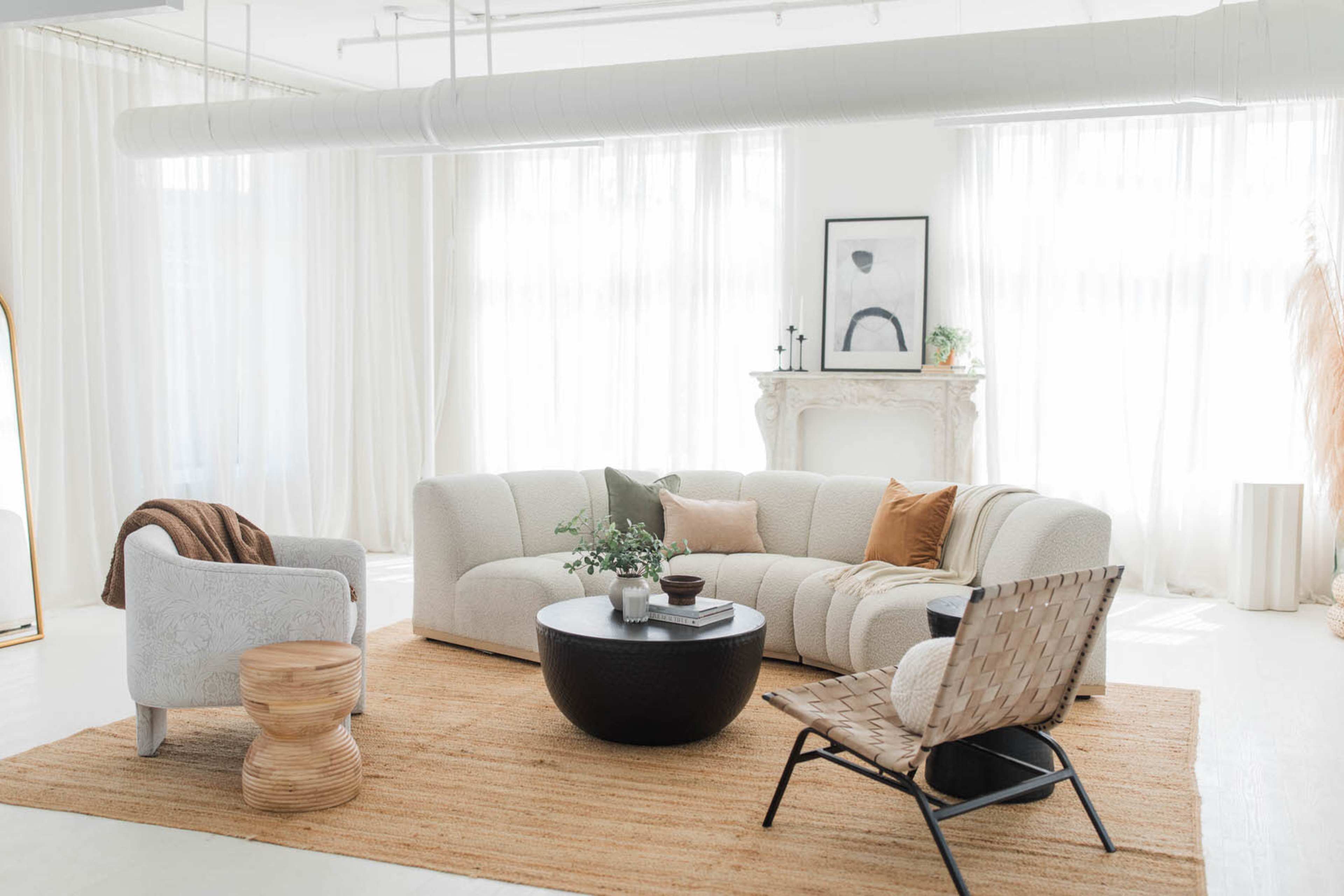 A modern living room features a light-colored sectional sofa, a round coffee table, and various seating options arranged on a natural fiber rug under large windows.