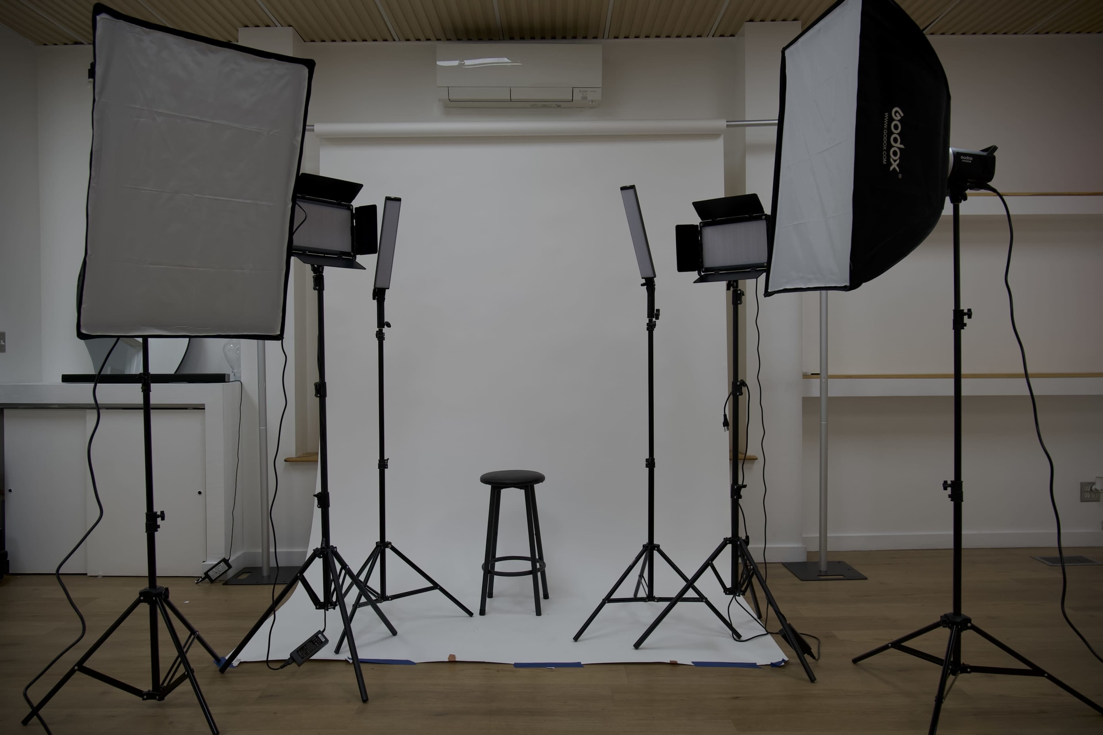 A photography setup features a stool in front of a white backdrop, surrounded by studio lights and softboxes.