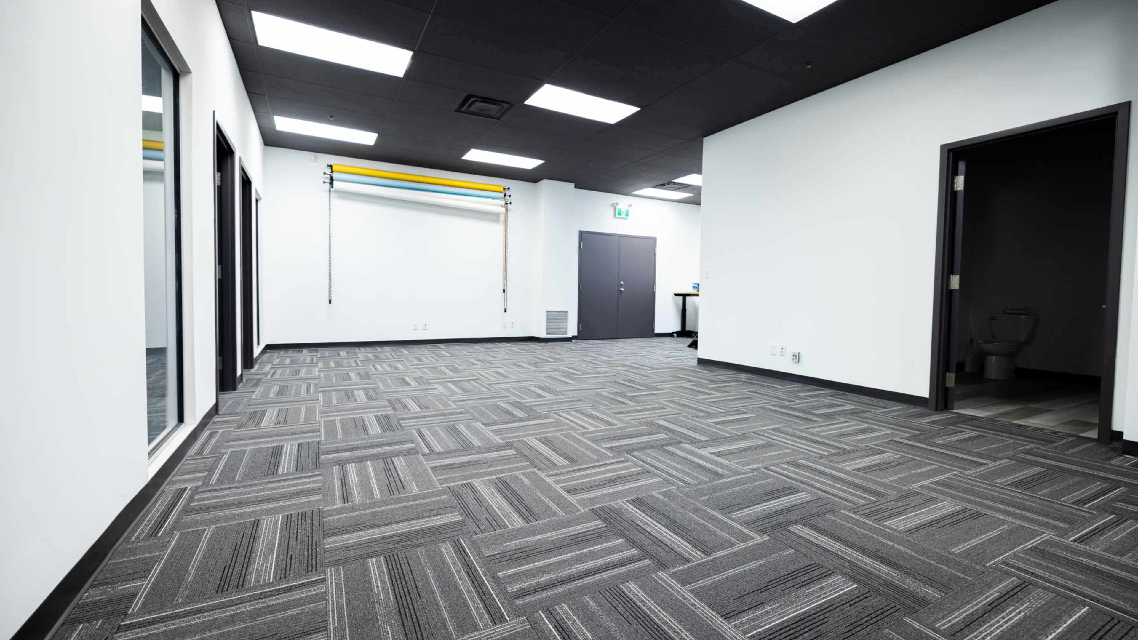 A large, empty room with gray carpeting, white walls, and overhead fluorescent lighting, featuring mirrors and a closed door on one side.