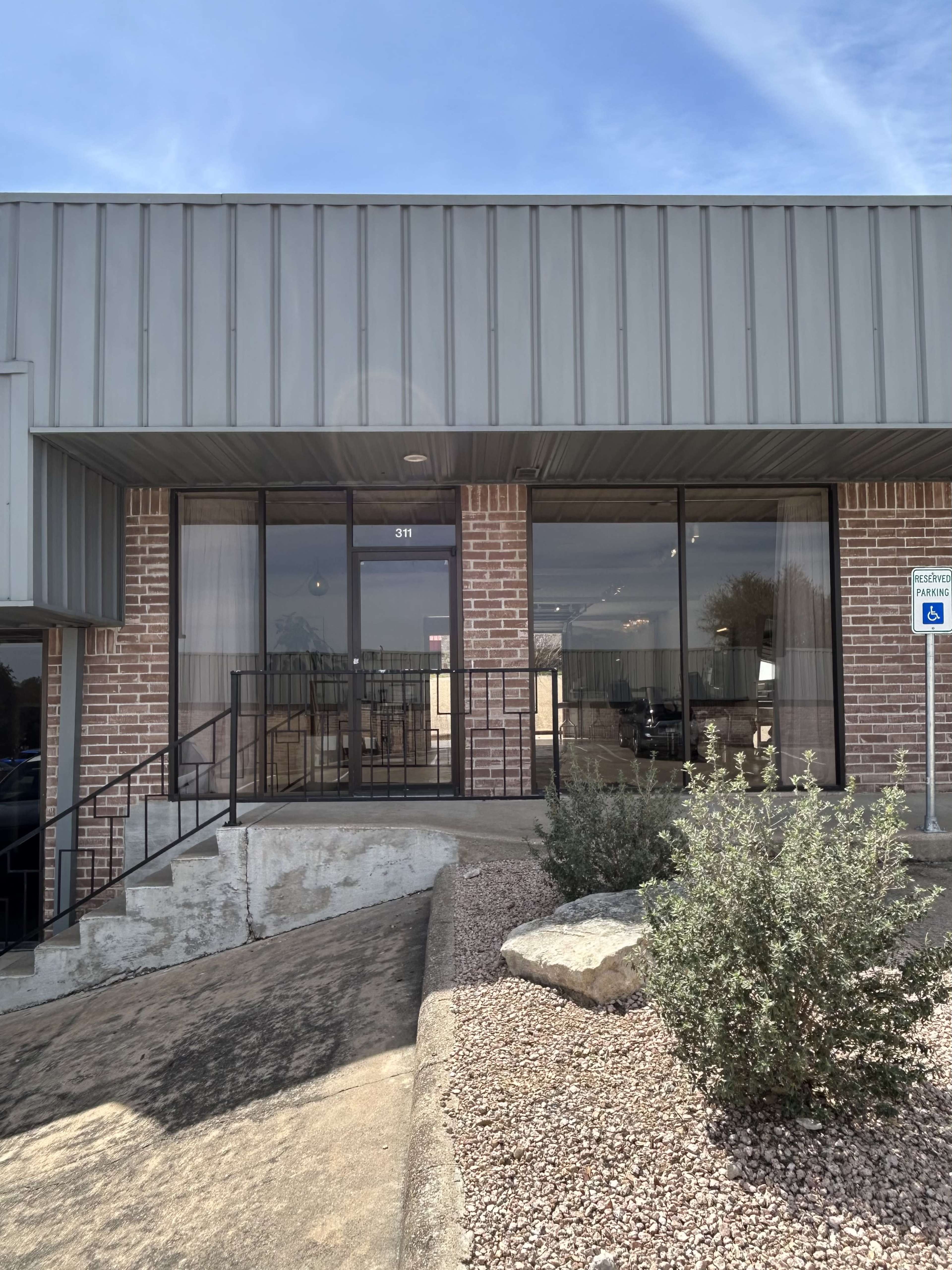 The image shows the exterior of a commercial building with a brick facade and large glass windows, featuring an entrance with a wheelchair ramp.