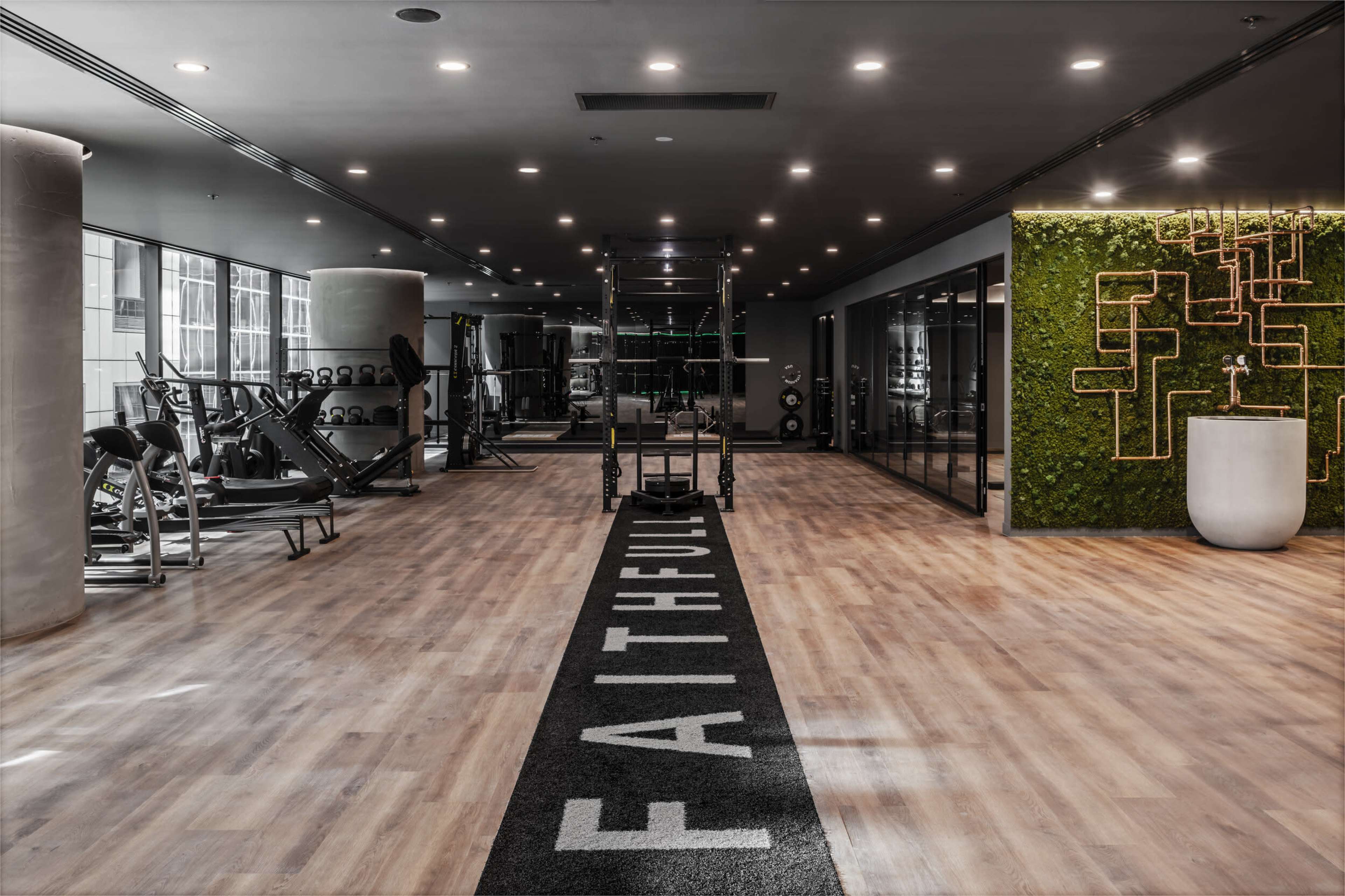 A modern gym features wooden flooring, workout equipment on one side, and a wall adorned with greenery and a fitness area.