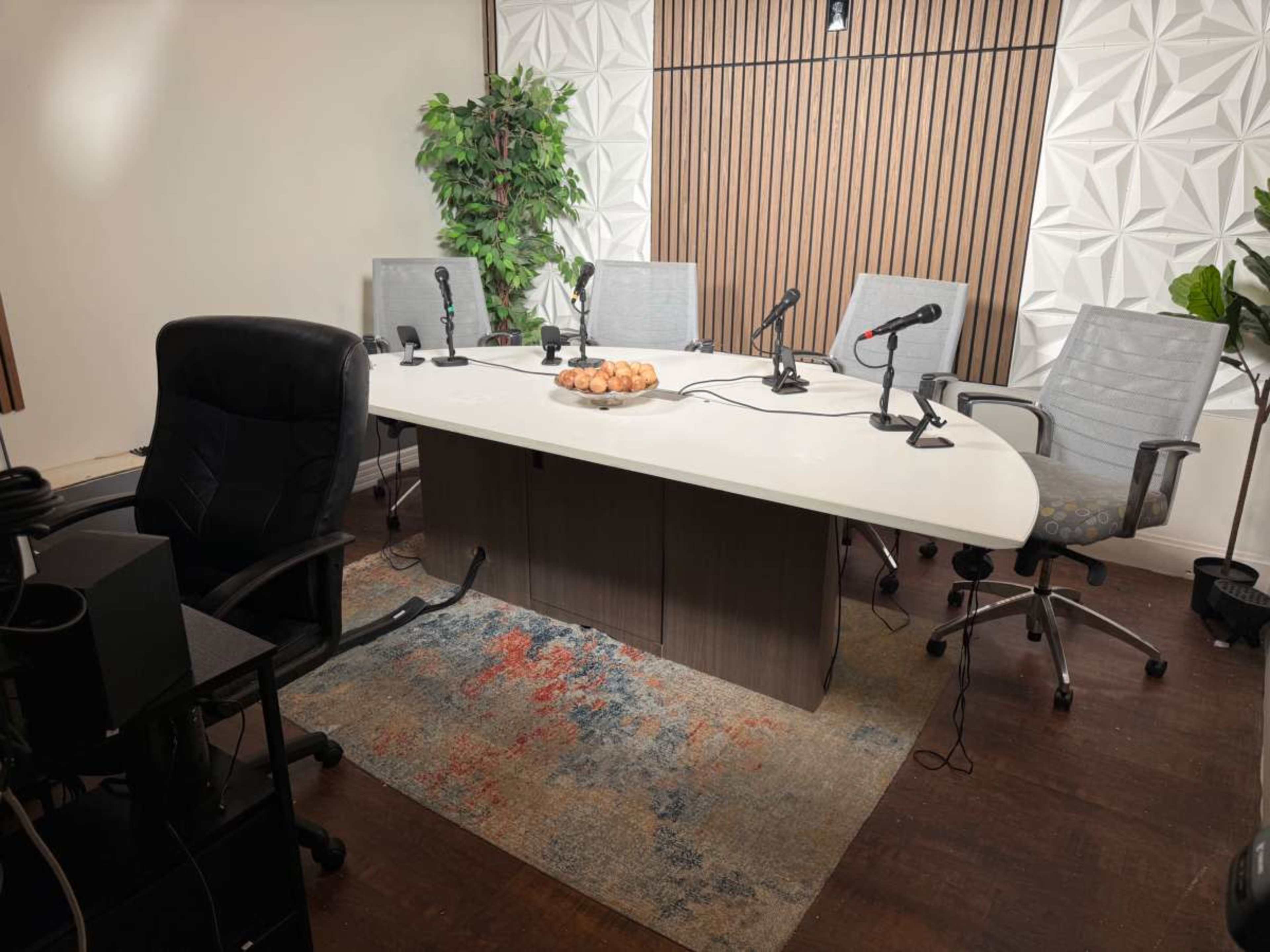 A conference room is set up with a large white table, six chairs, and several microphones, with a decorative plant and wall patterns in the background.