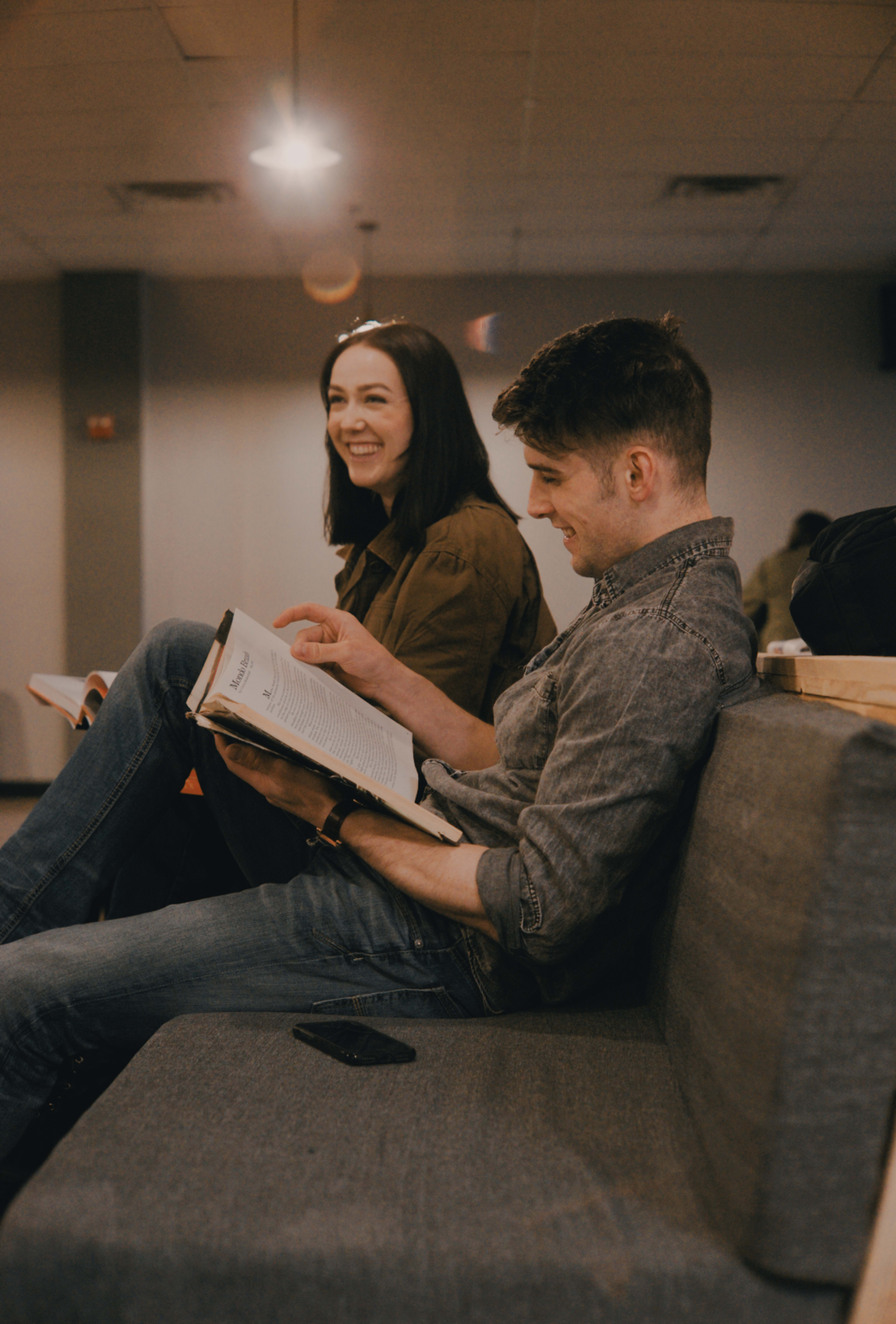 A man and woman sit on a couch, smiling and reading books in a well-lit indoor space.