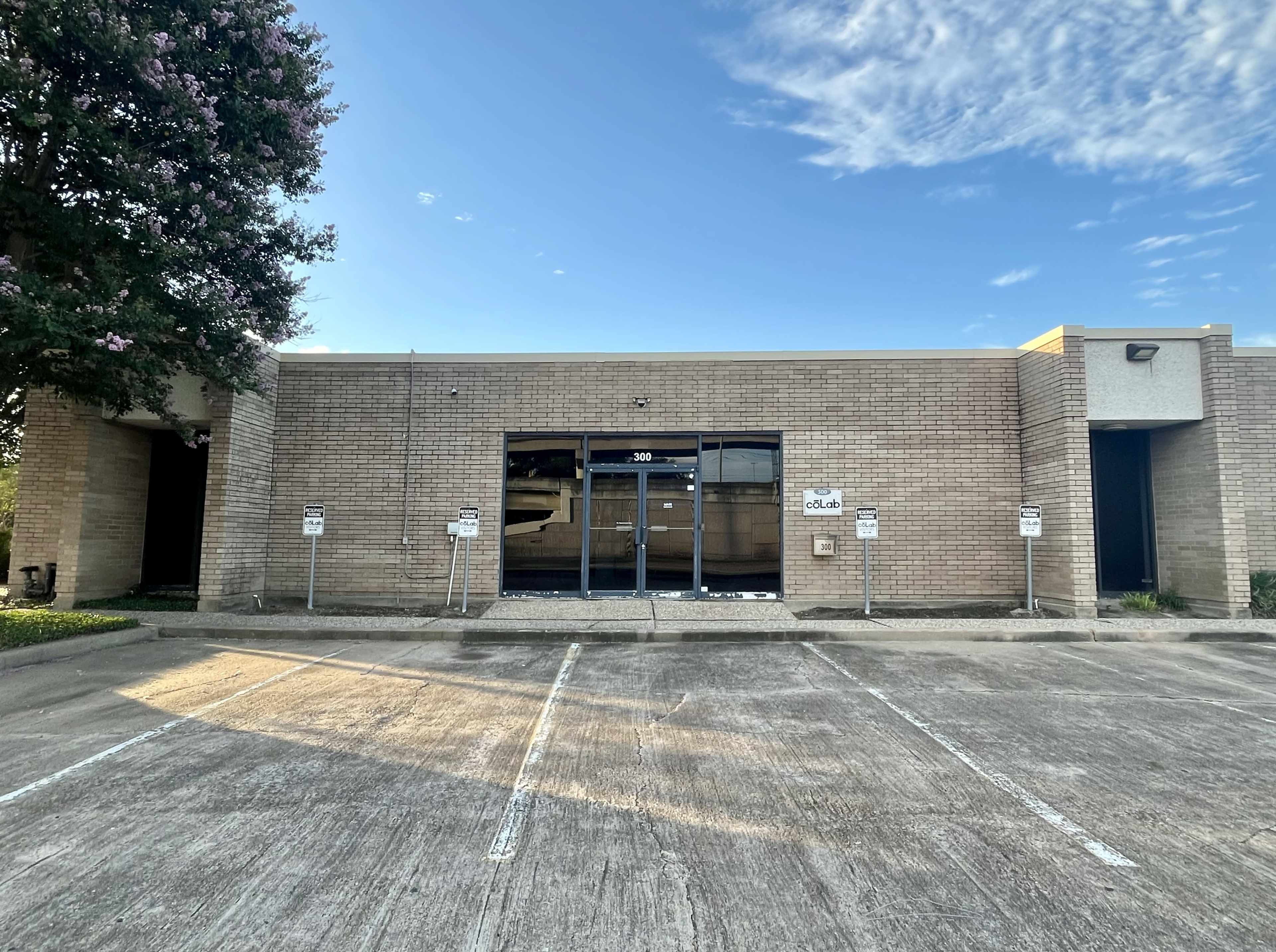 The image shows a single-story commercial building with large glass windows and a parking lot in front, featuring several empty parking spaces.