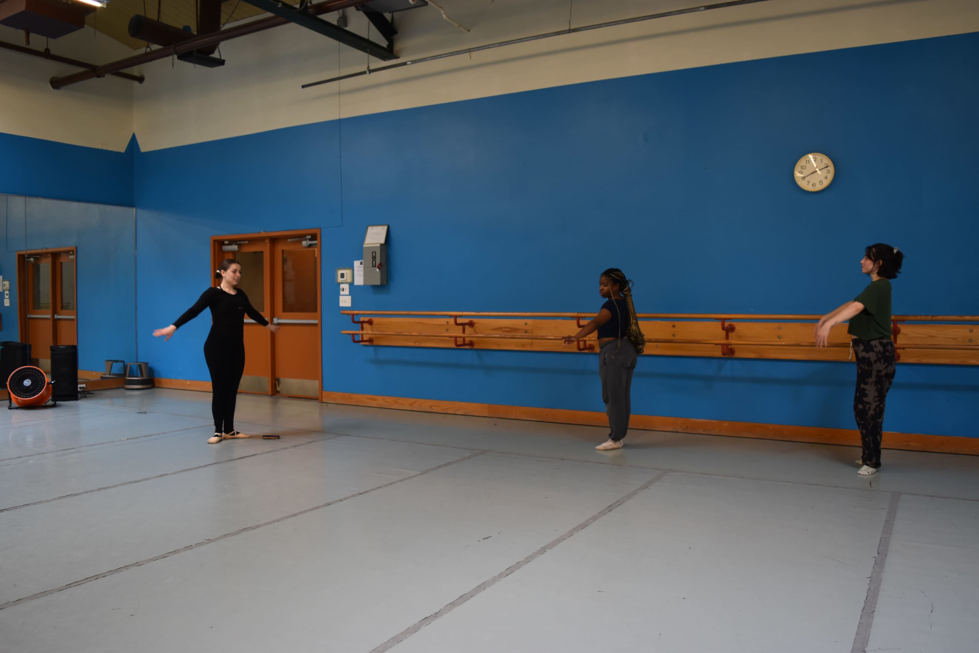 Three dancers practice in a studio with mirrors and a blue wall, each positioned near a wooden ballet bar.