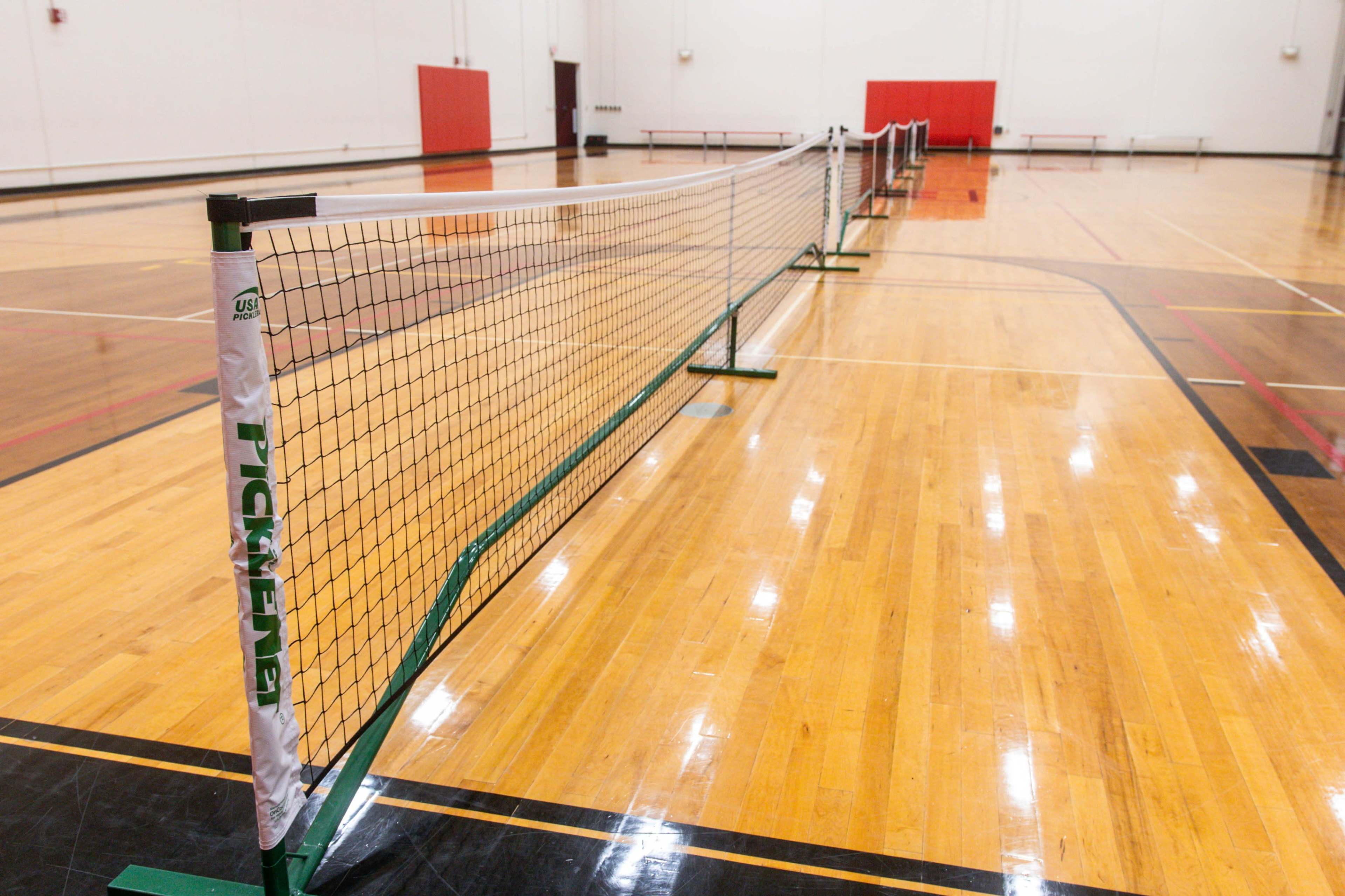 The image shows multiple tennis nets set up in a gymnasium with polished wooden floors and red walls.