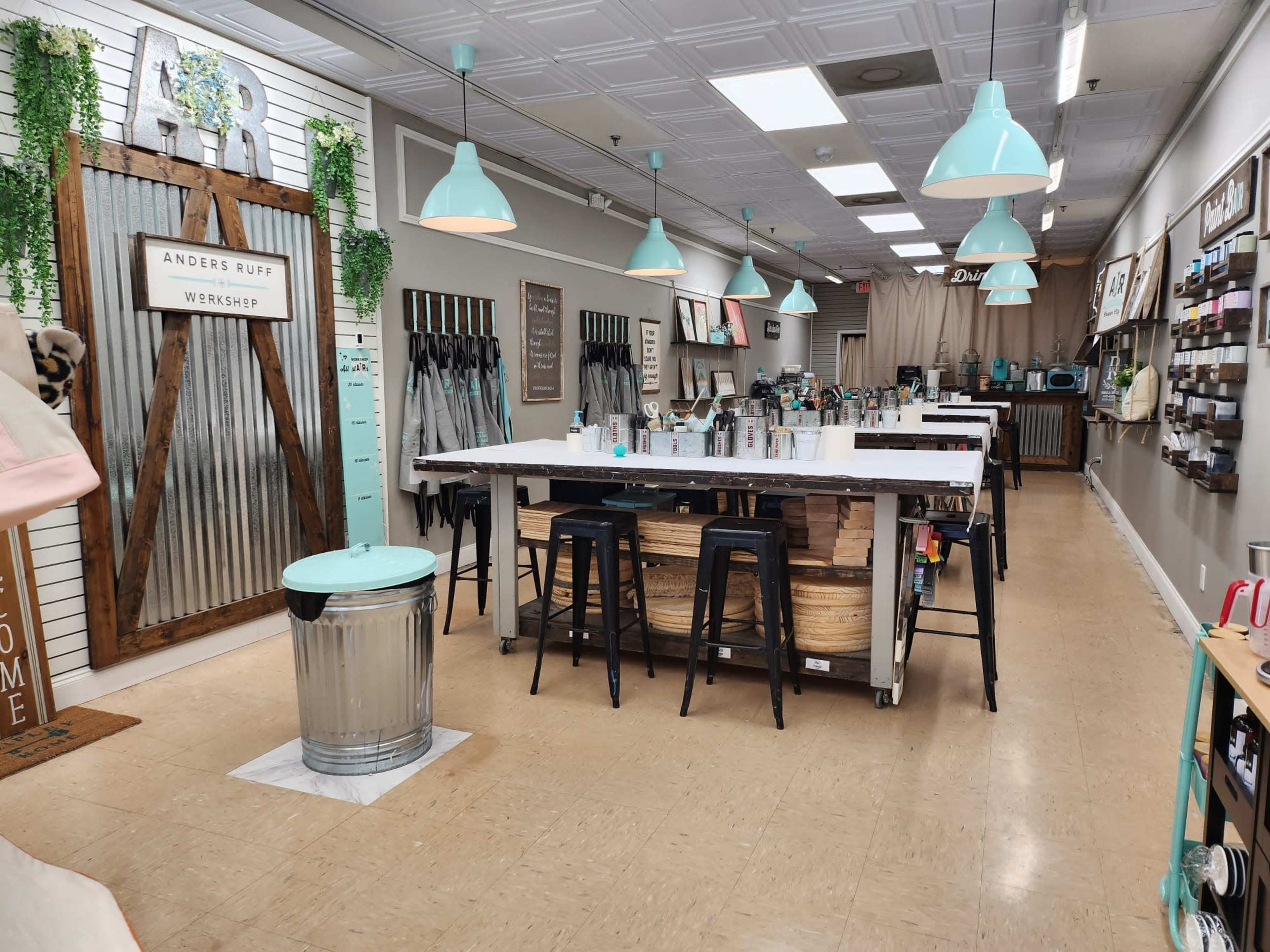 A spacious workshop with a long white counter, black stools, and various crafting supplies organized along the walls.