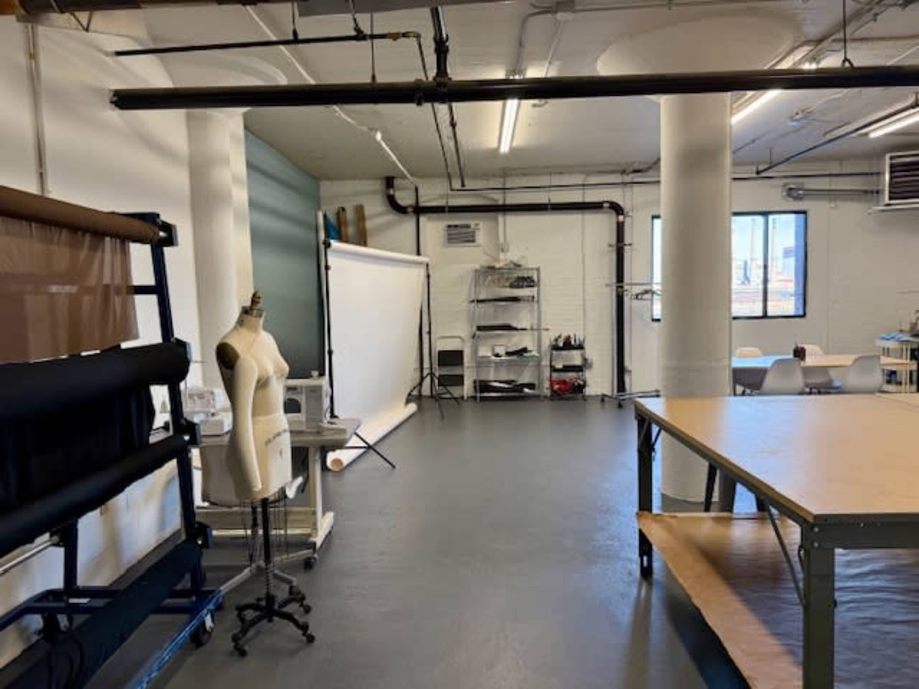 The image shows a spacious studio with white walls, large columns, a backdrop setup, a mannequin, and tables for fabric work.