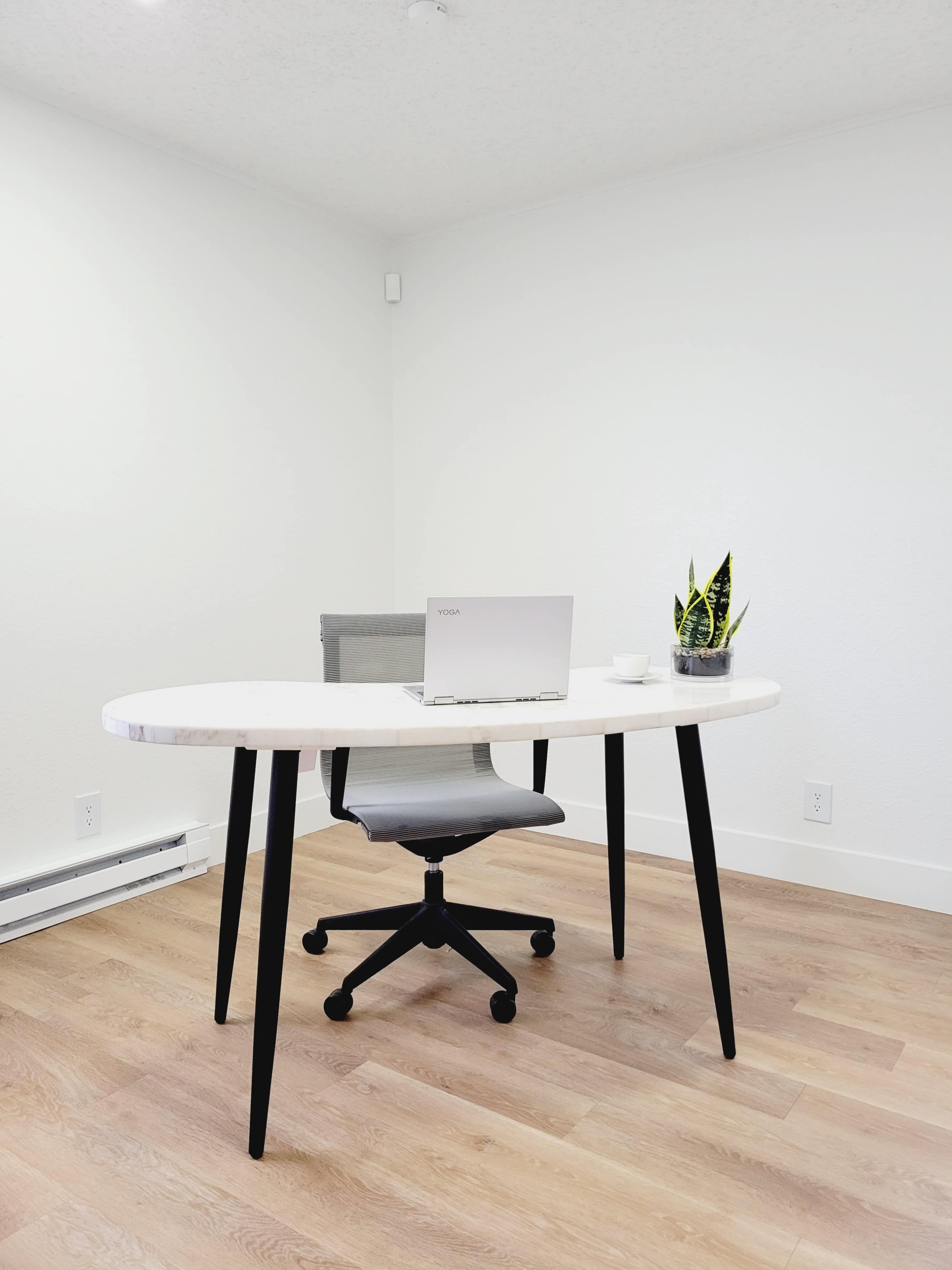 A modern workspace features a round white table, a gray office chair, a laptop, and a small plant on a light wooden floor.
