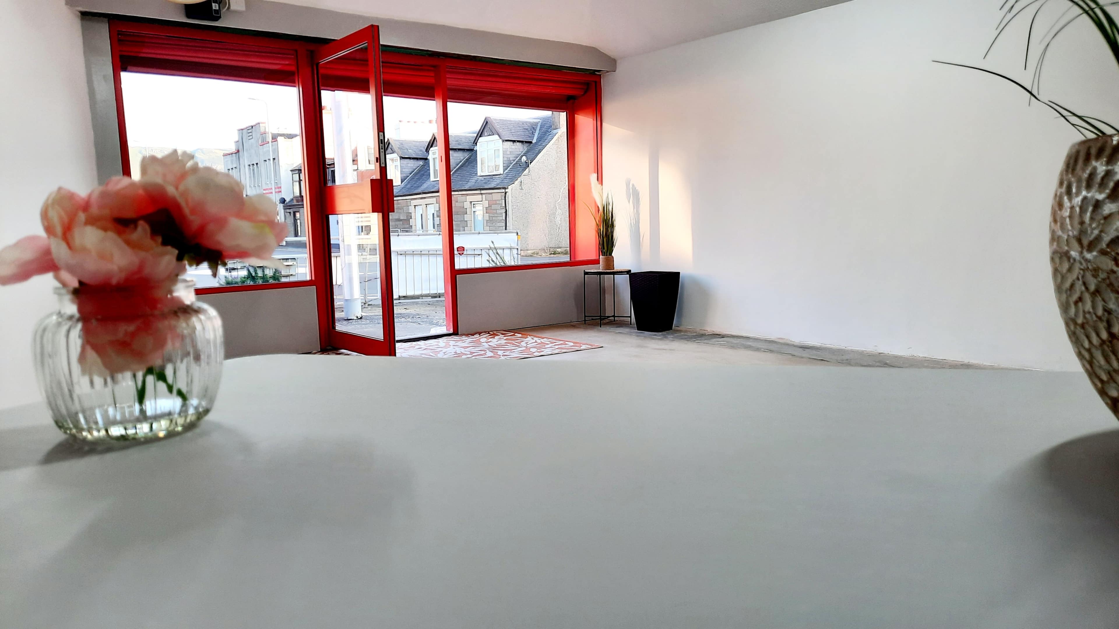 A bright, minimalist room features a red-framed window overlooking rooftops, with a glass vase of flowers and a decorative plant in the foreground.