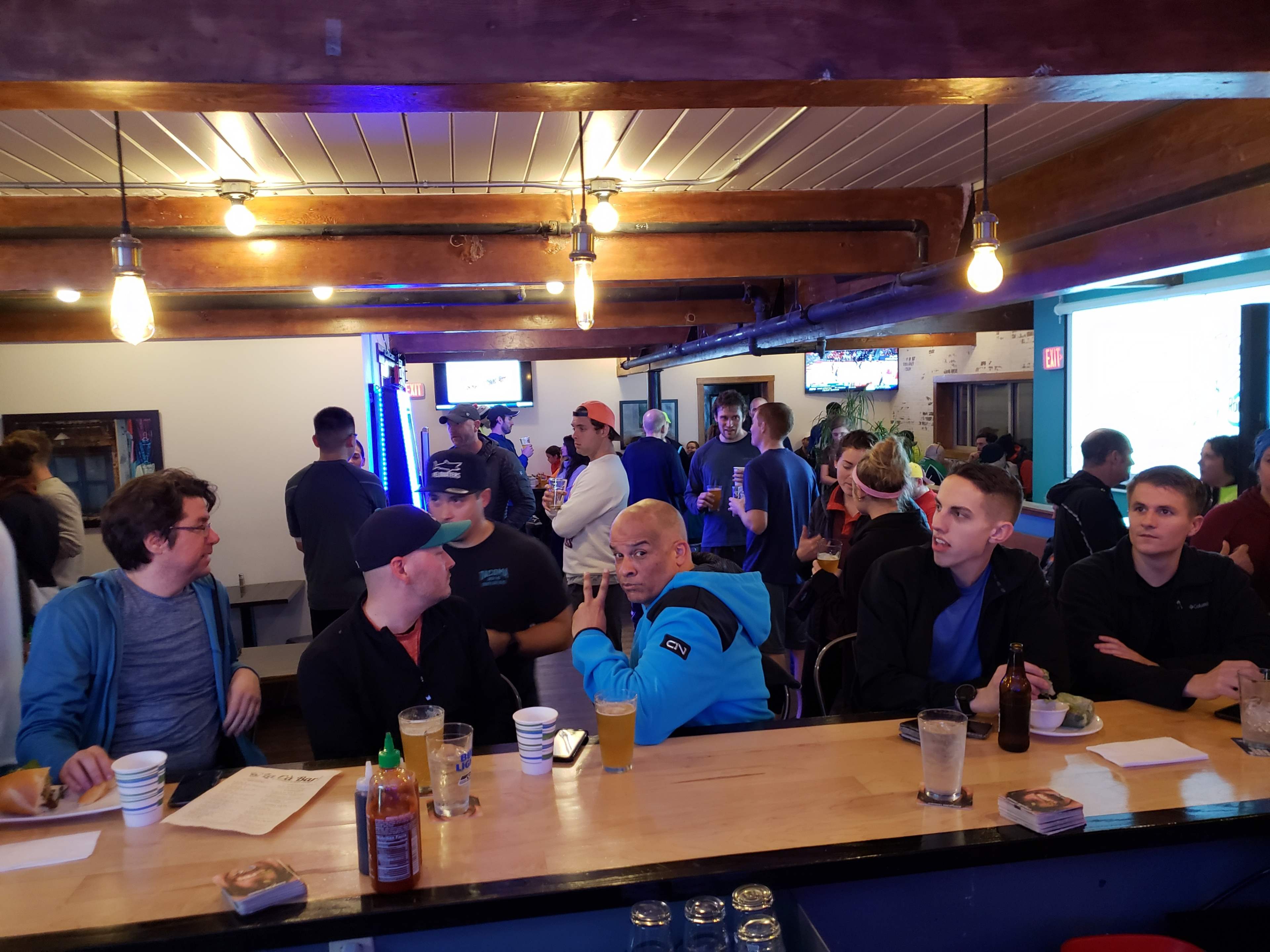 A group of people are seated at a bar with a lively atmosphere, while others socialize in the background.