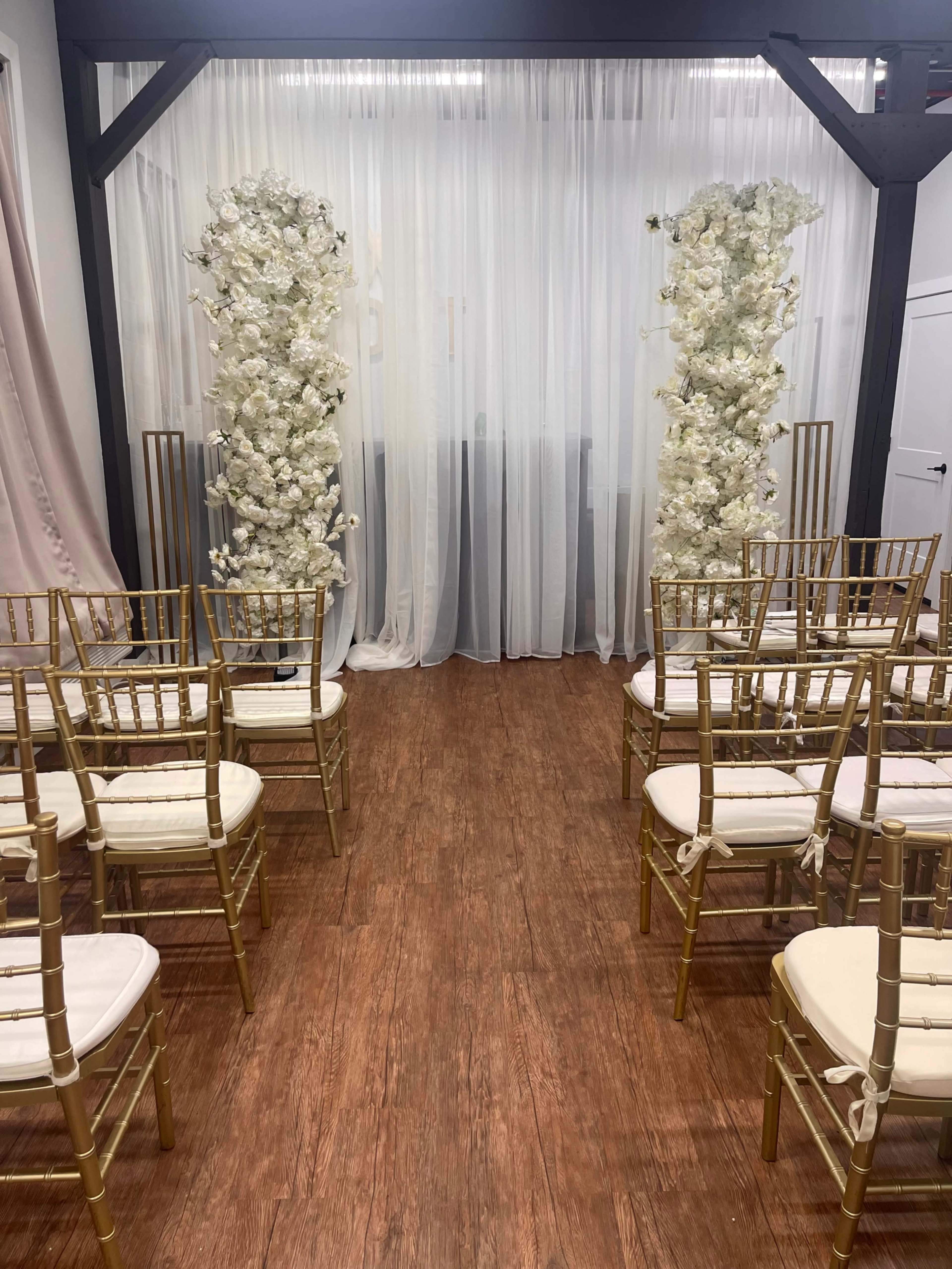 The image shows a wedding ceremony setup with two floral arrangements and rows of gold chairs facing a draped backdrop.