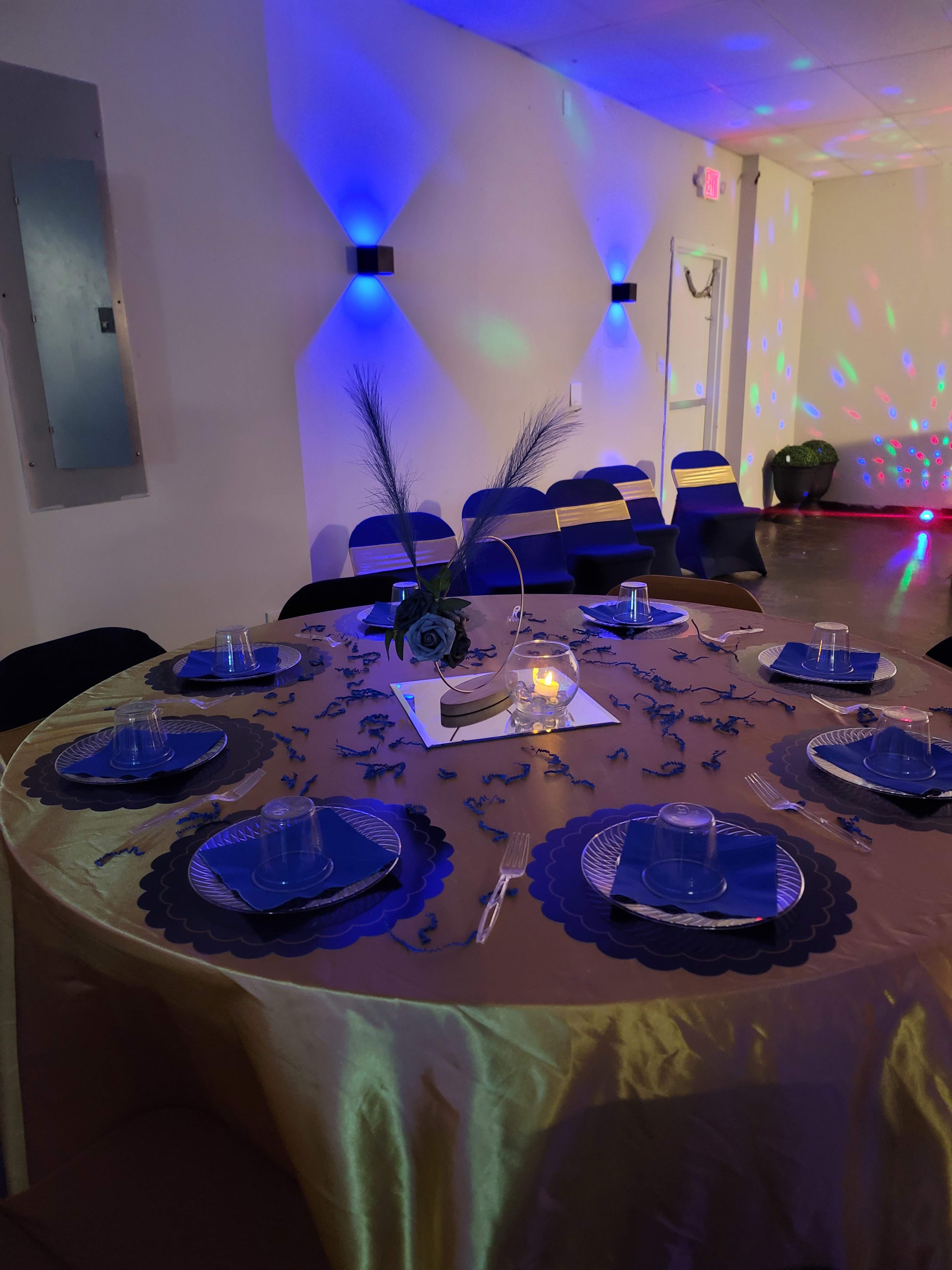 A round table is set with blue plates and clear cups, surrounded by chairs, and decorated with a candle and flower centerpiece, with colored lights casting patterns on the walls.