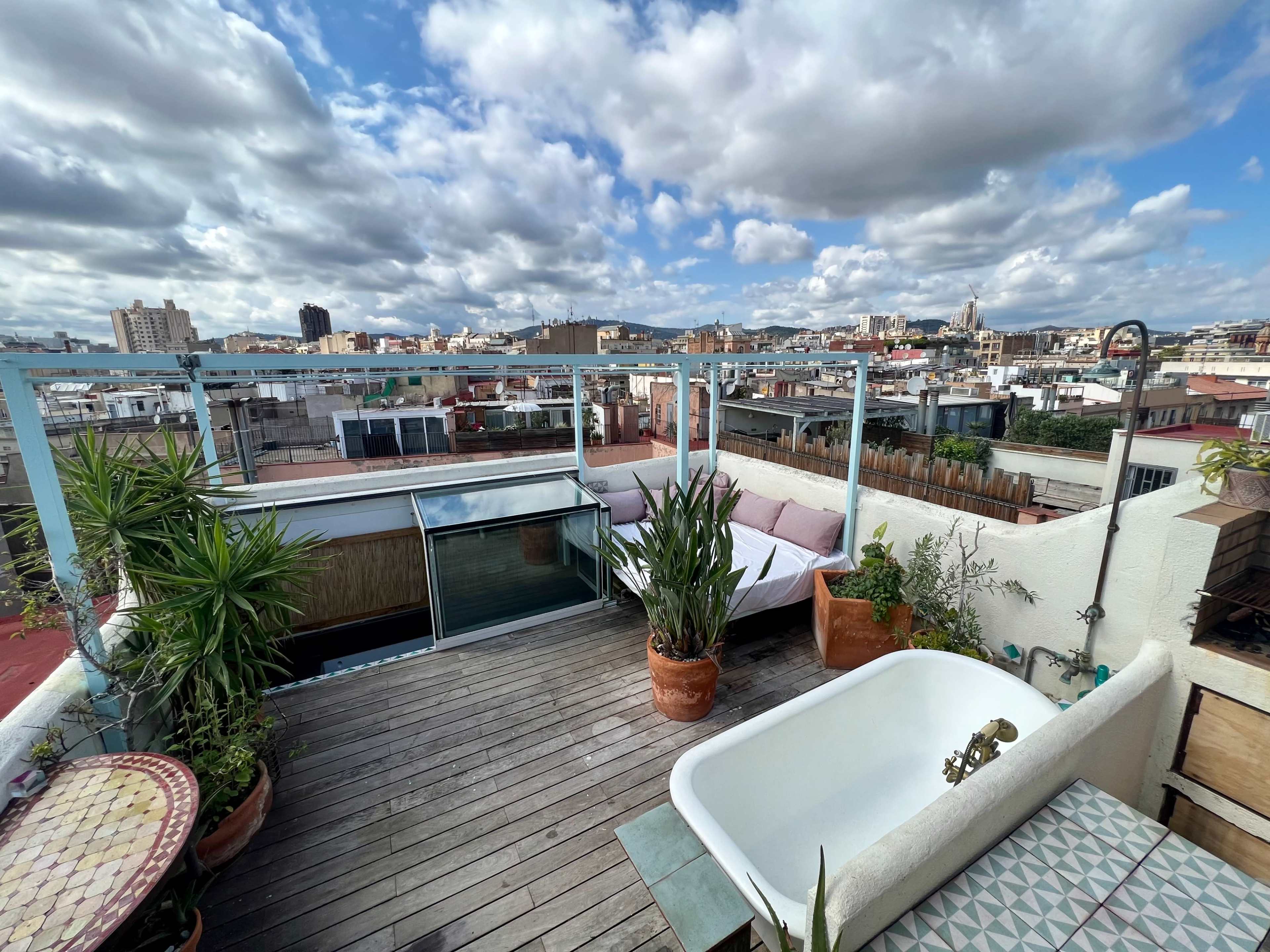 The image shows a rooftop terrace with a wooden deck, potted plants, a bathtub, and a view of a city skyline under a partly cloudy sky.