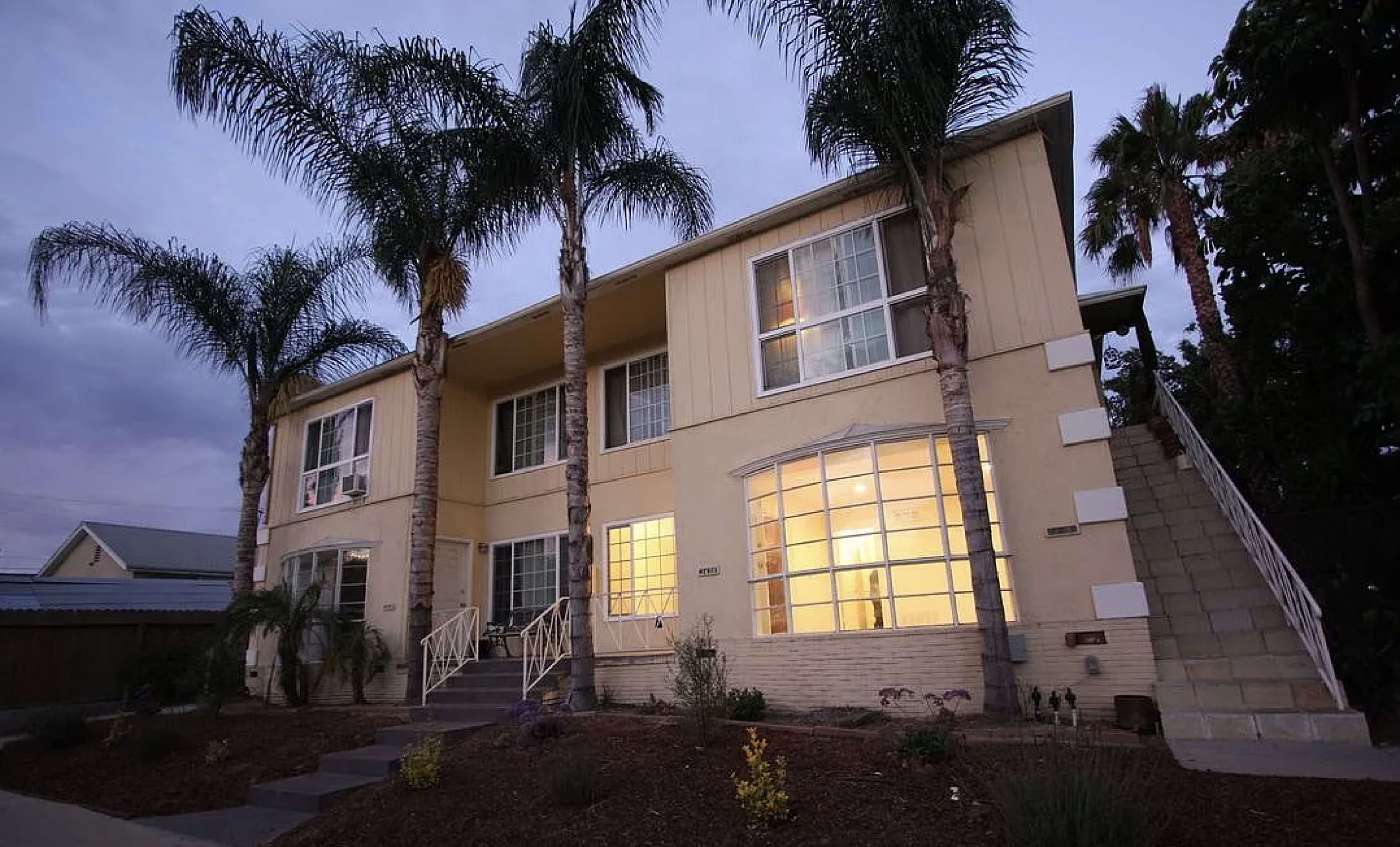 The image shows a two-story residential building with palm trees in front and warm light emanating from the windows.