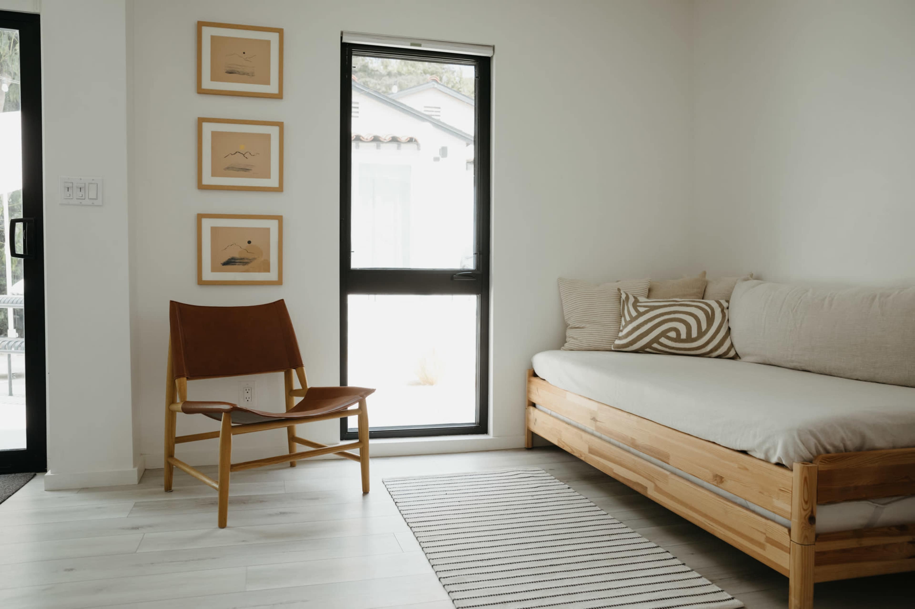 The scene features a minimalist living space with a wooden daybed, a brown leather chair, and framed artwork on the wall beside a large window.