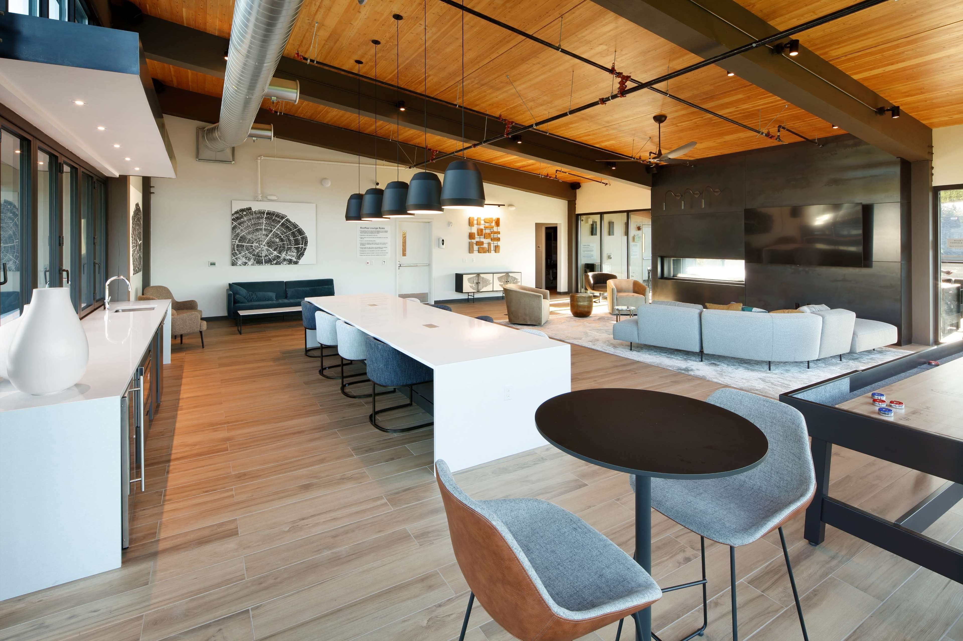 A modern open-plan living space with a kitchen, dining area, and lounge, featuring large windows and wooden ceiling beams.