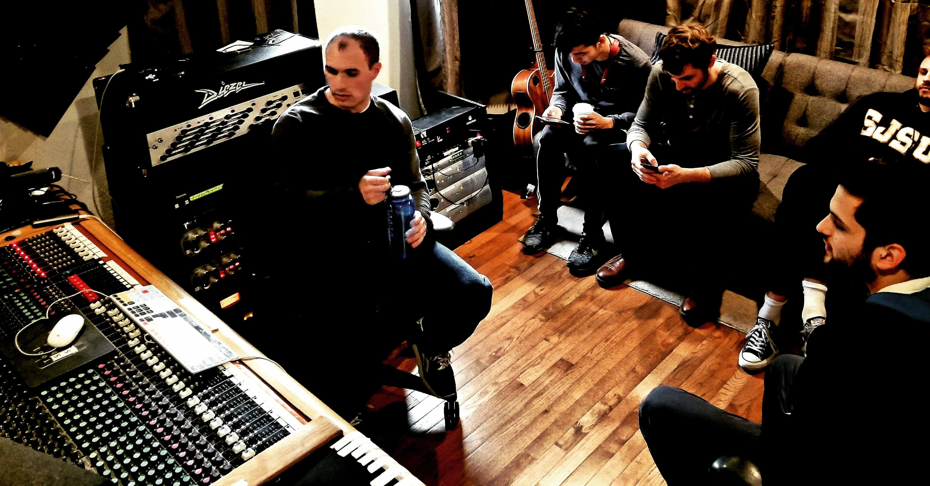 A group of four men sits in a recording studio, with one man speaking while others listen and engage with their phones.