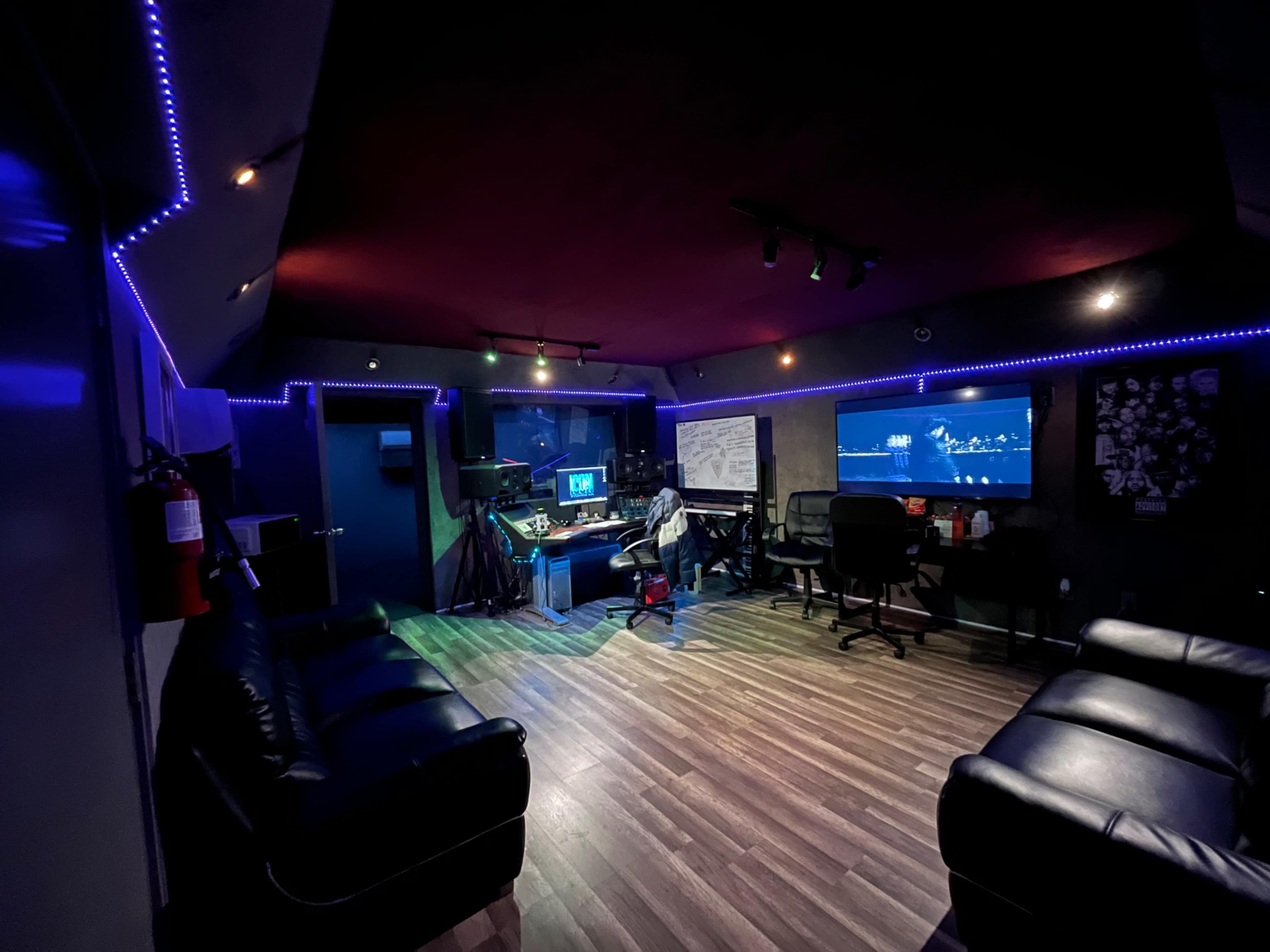 A recording studio features a central workspace with computers and sound equipment, surrounded by black couches and illuminated by colored LED lights.