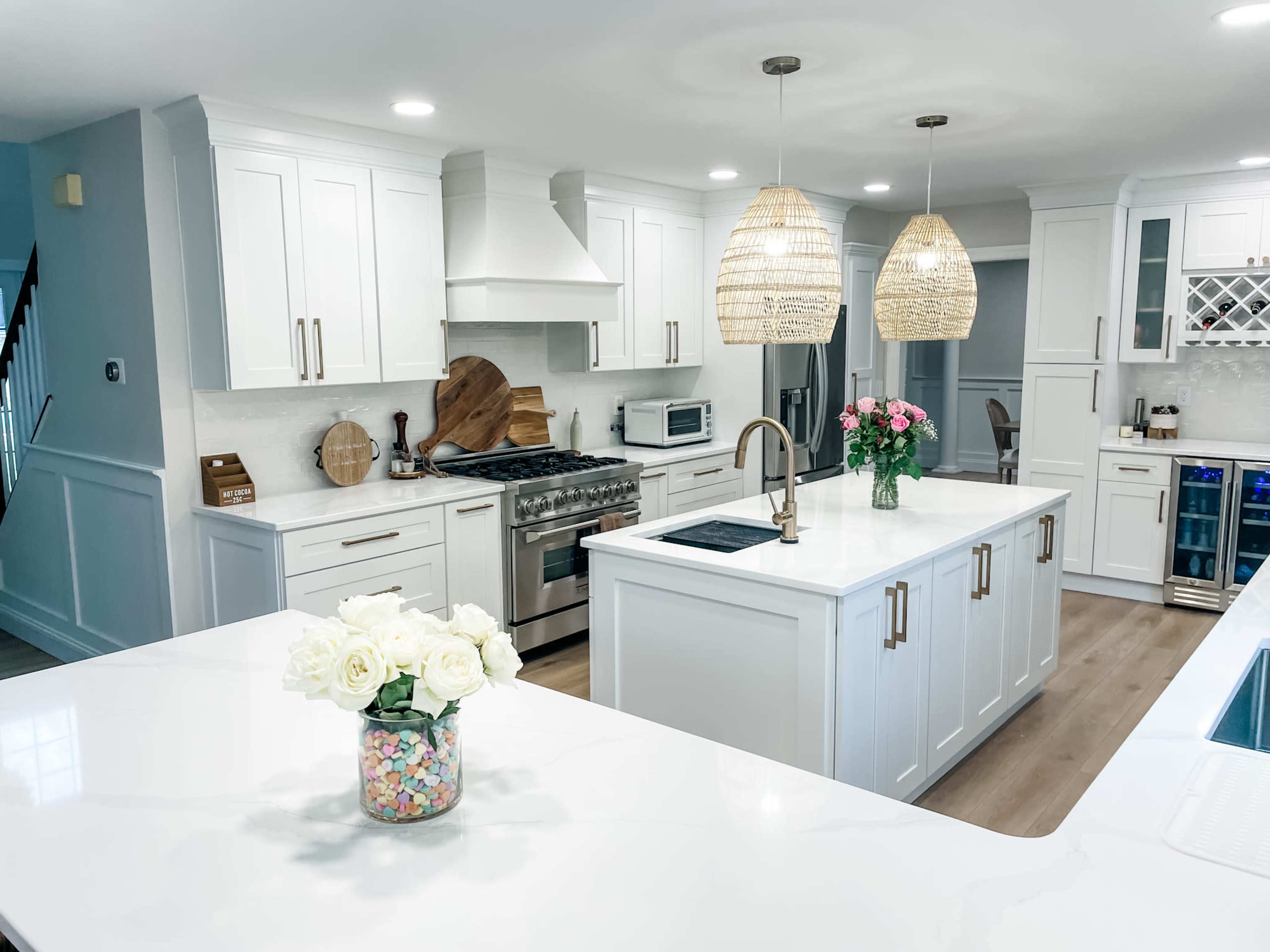 A modern kitchen features white cabinetry, a large island with a sink, and pendant lighting above, complemented by freshly cut flowers in a jar on the counter.
