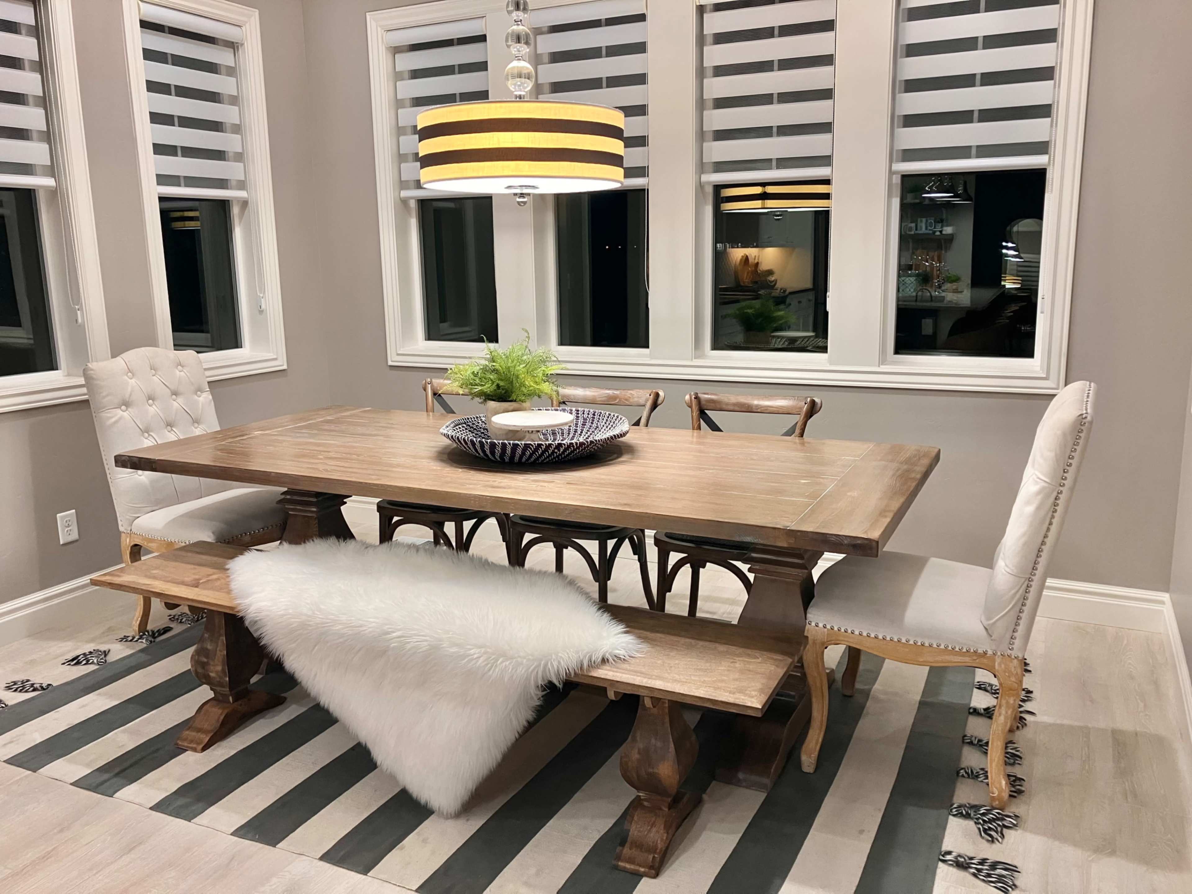 A wooden dining table surrounded by chairs is set in a well-lit dining area with large windows and a pendant light above.
