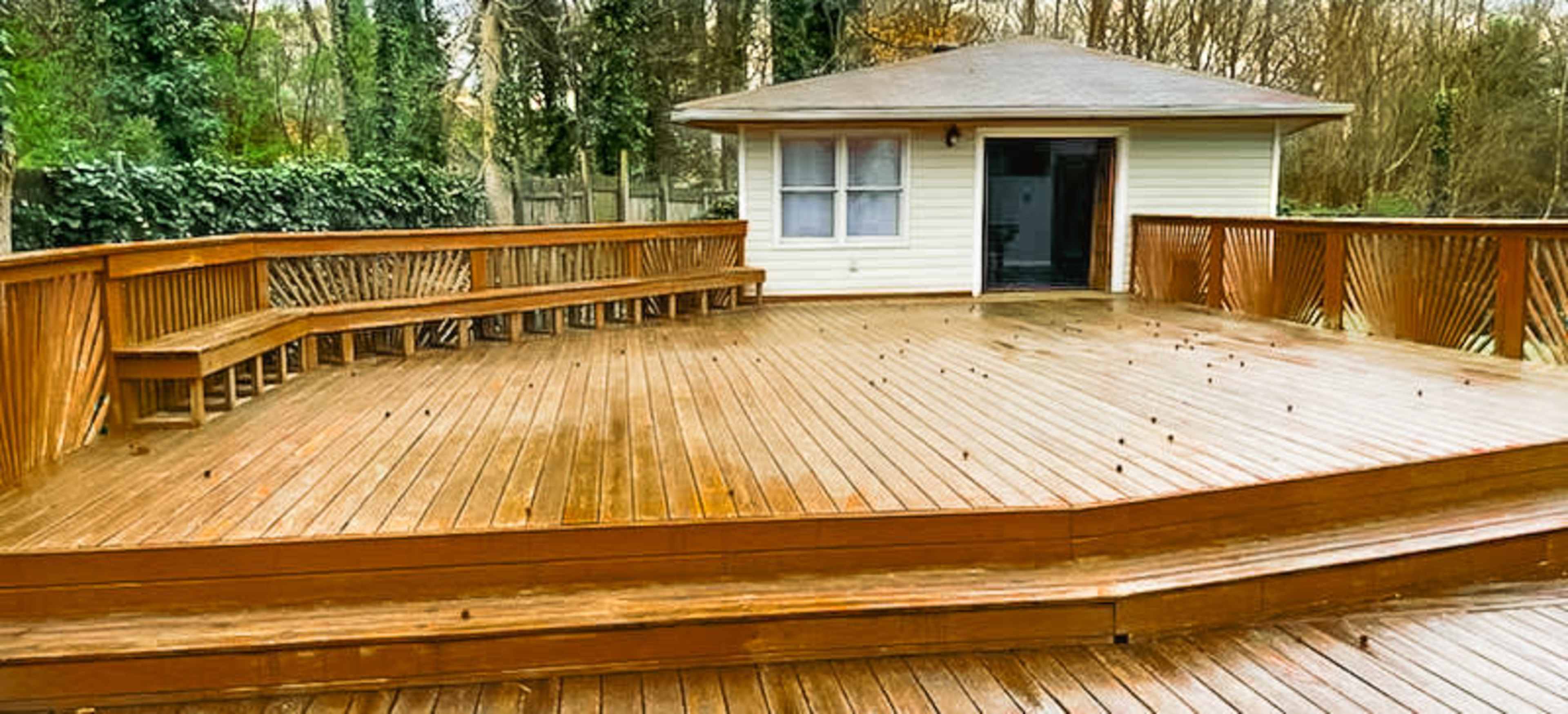 A large wooden deck in front of a small house, featuring benches along the edge and surrounded by trees.
