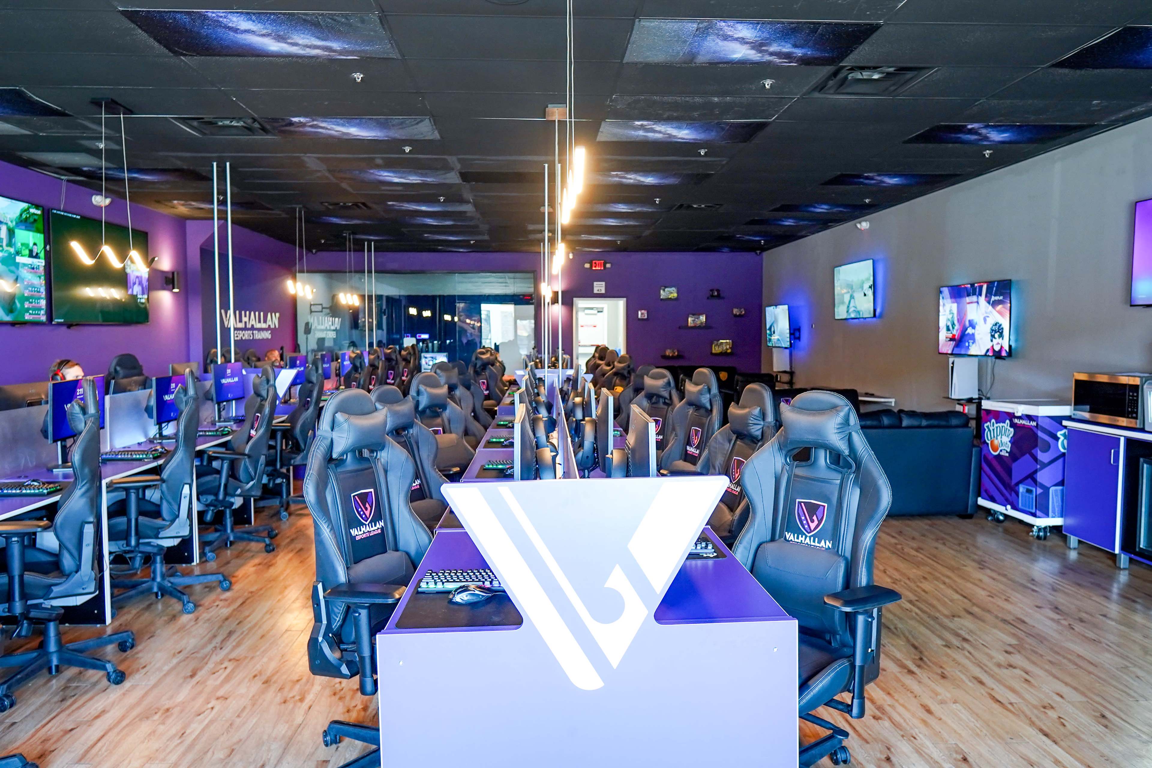 The image shows a spacious esports gaming lounge equipped with rows of gaming chairs and computers, featuring a purple color scheme and several screens displaying various games.