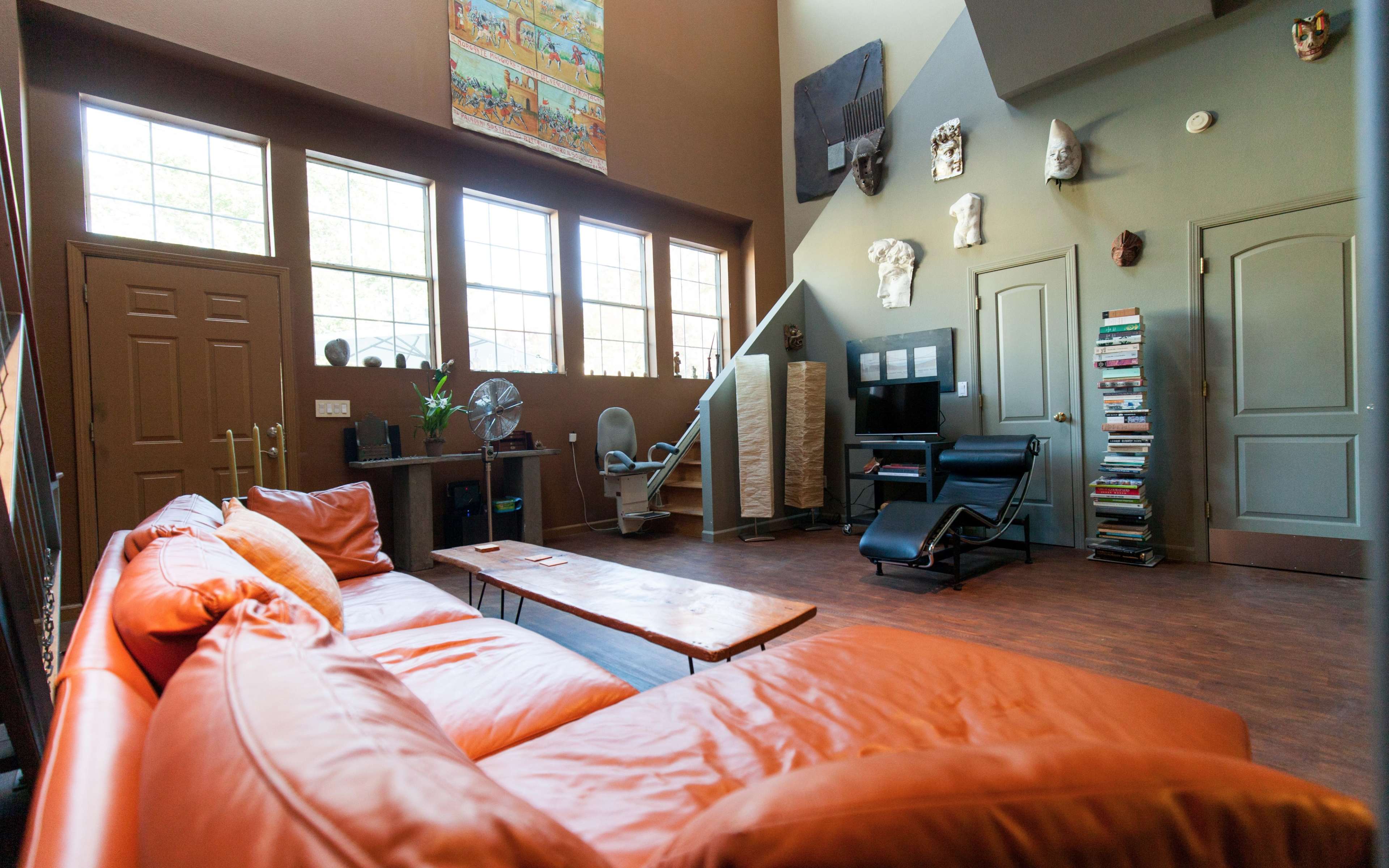 The interior of a living room features an orange sectional sofa, a wooden coffee table, and a corner with a computer desk, surrounded by windows and decorative wall art.