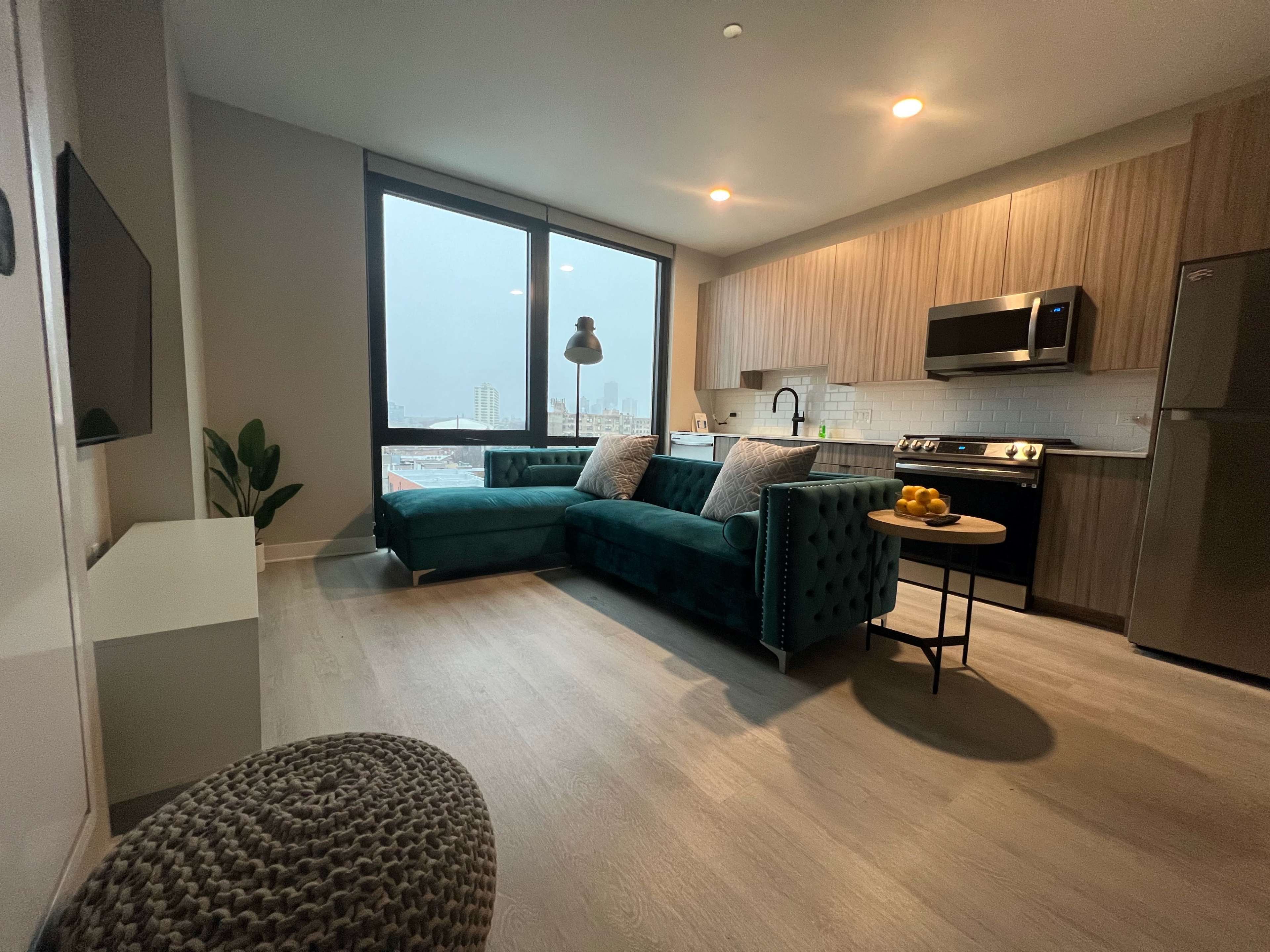 A modern living space features a green sectional couch, a kitchen area with wood cabinetry, and large windows overlooking a cityscape.
