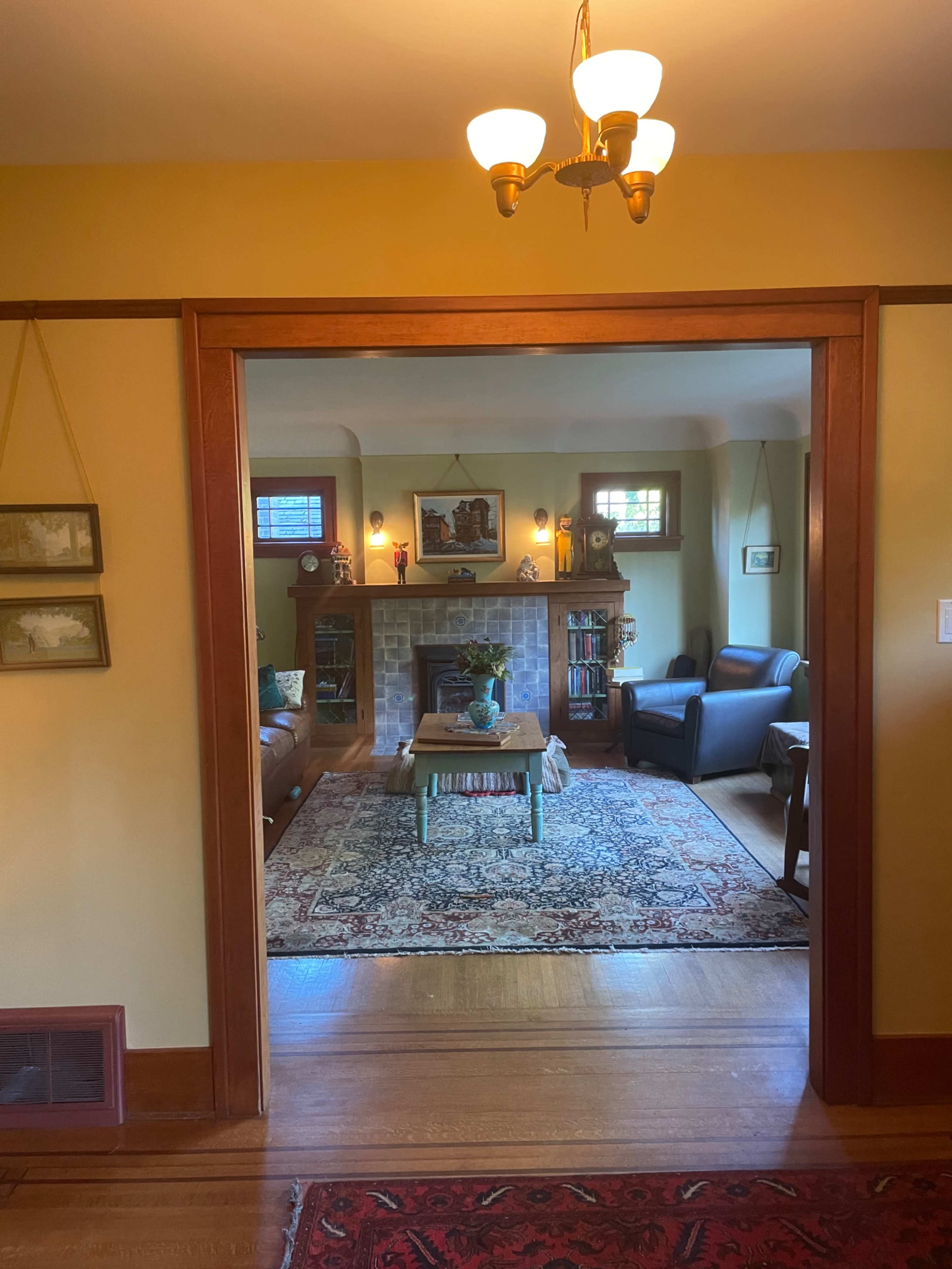 A cozy living room is framed by an open doorway, featuring a fireplace, a coffee table, and upholstered seating around a decorative rug.