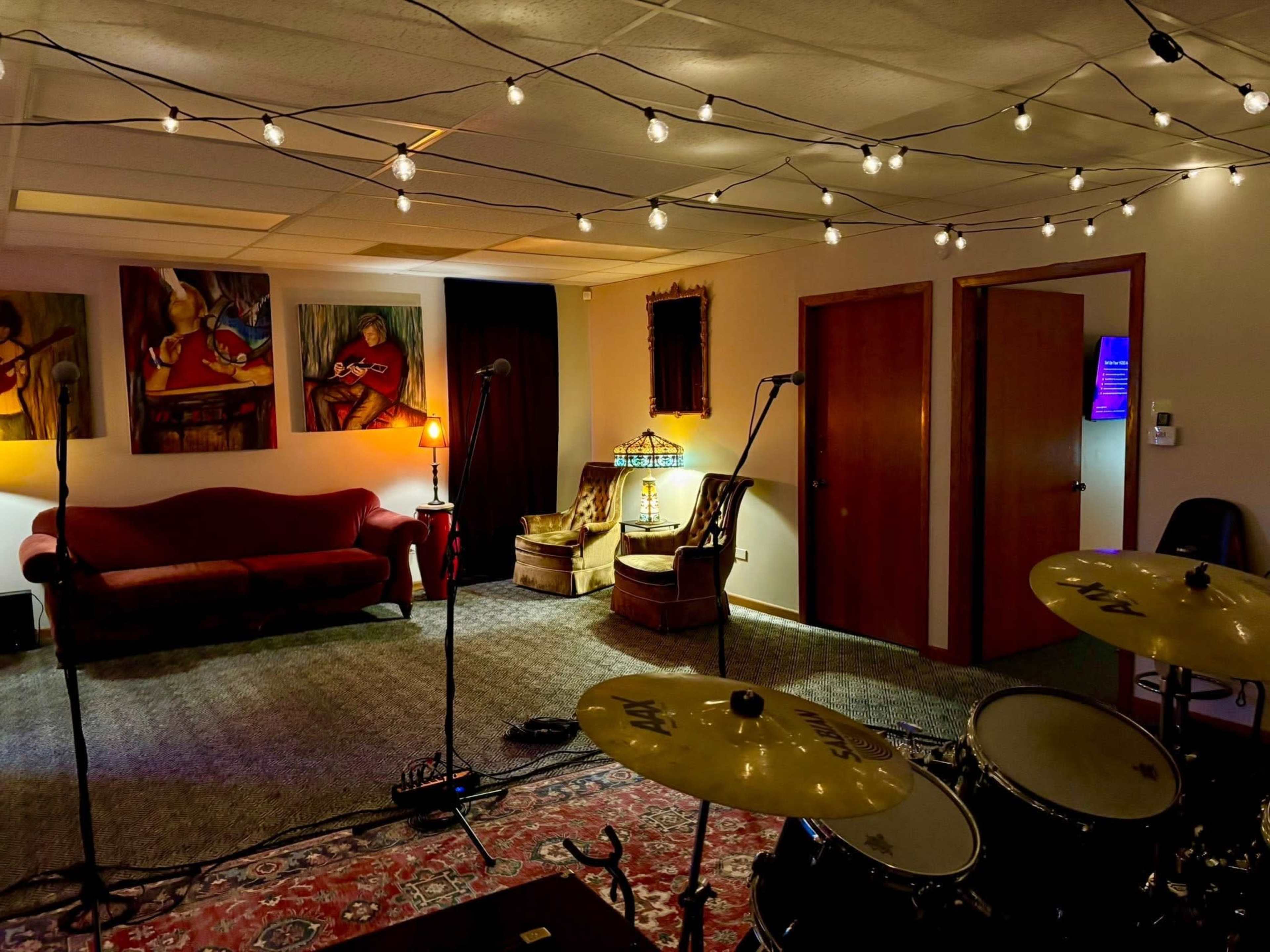 The image shows a music rehearsal space featuring a drum set, decorative lighting, and several artworks on the walls, along with a couch and chairs arranged in the room.