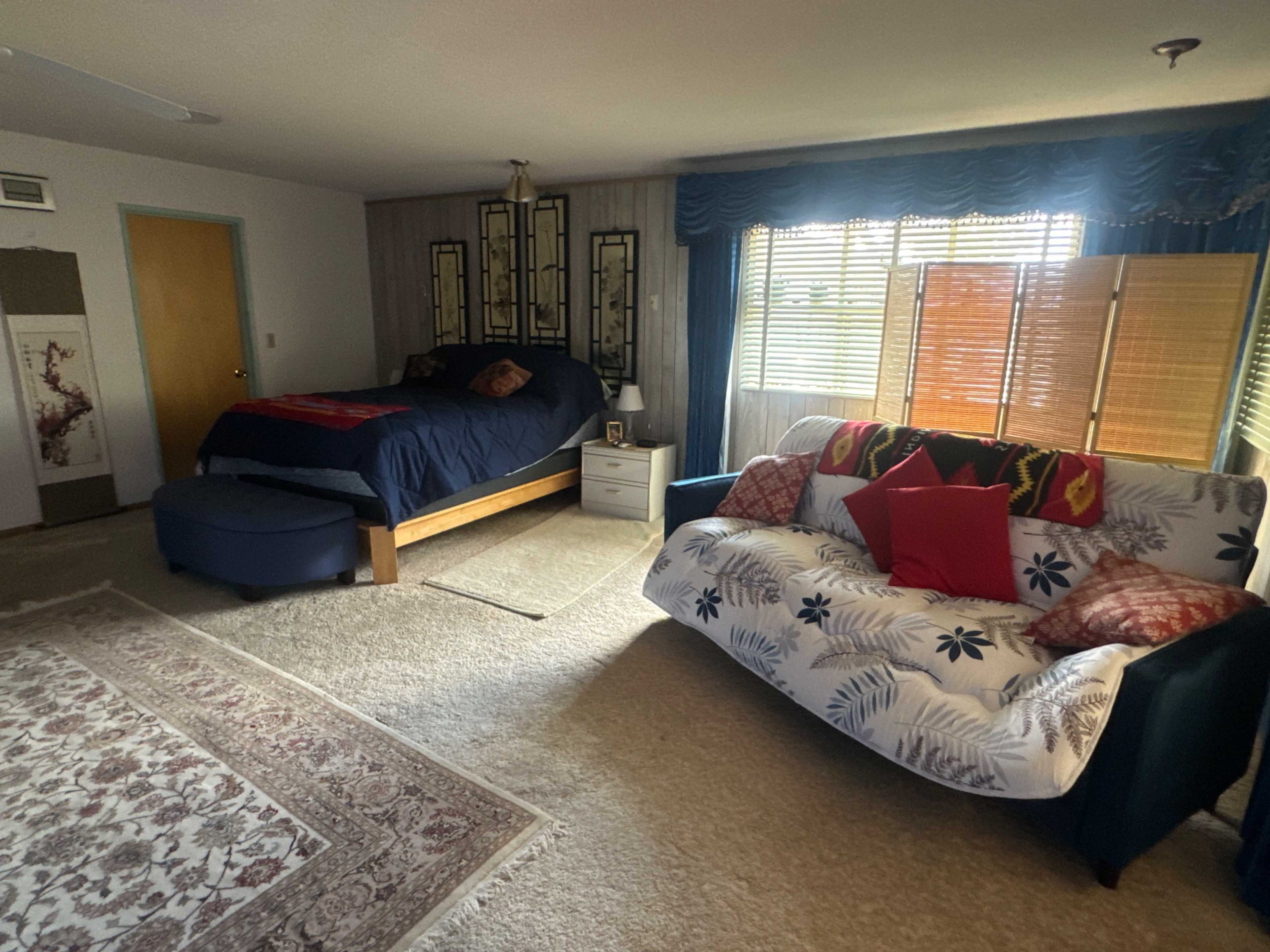 The image shows a cozy room with a bed on one side and a couch adorned with decorative pillows on the other, illuminated by natural light coming through window blinds.