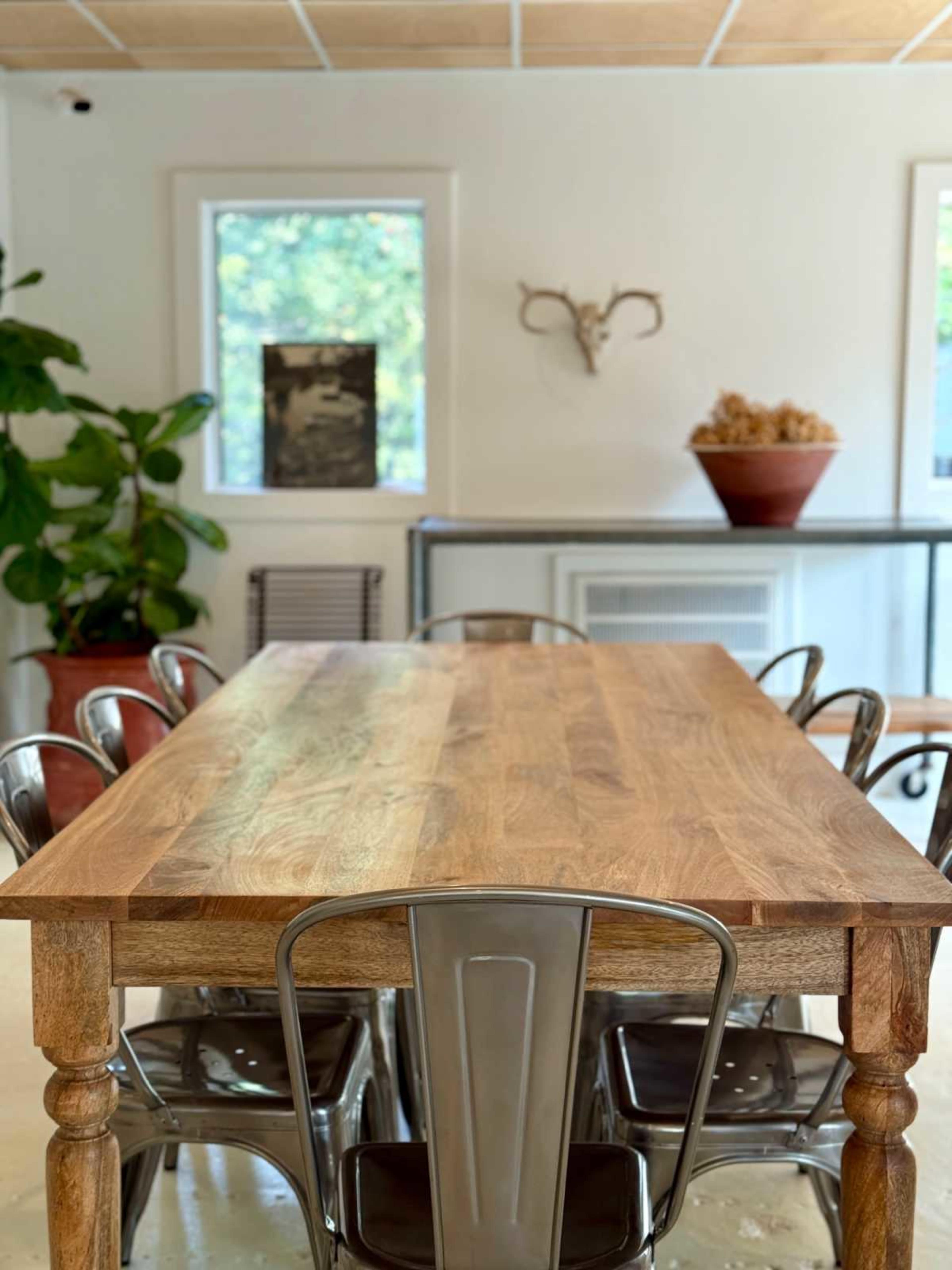A long wooden dining table with metal chairs sits in a bright room that features plants and wall decor.
