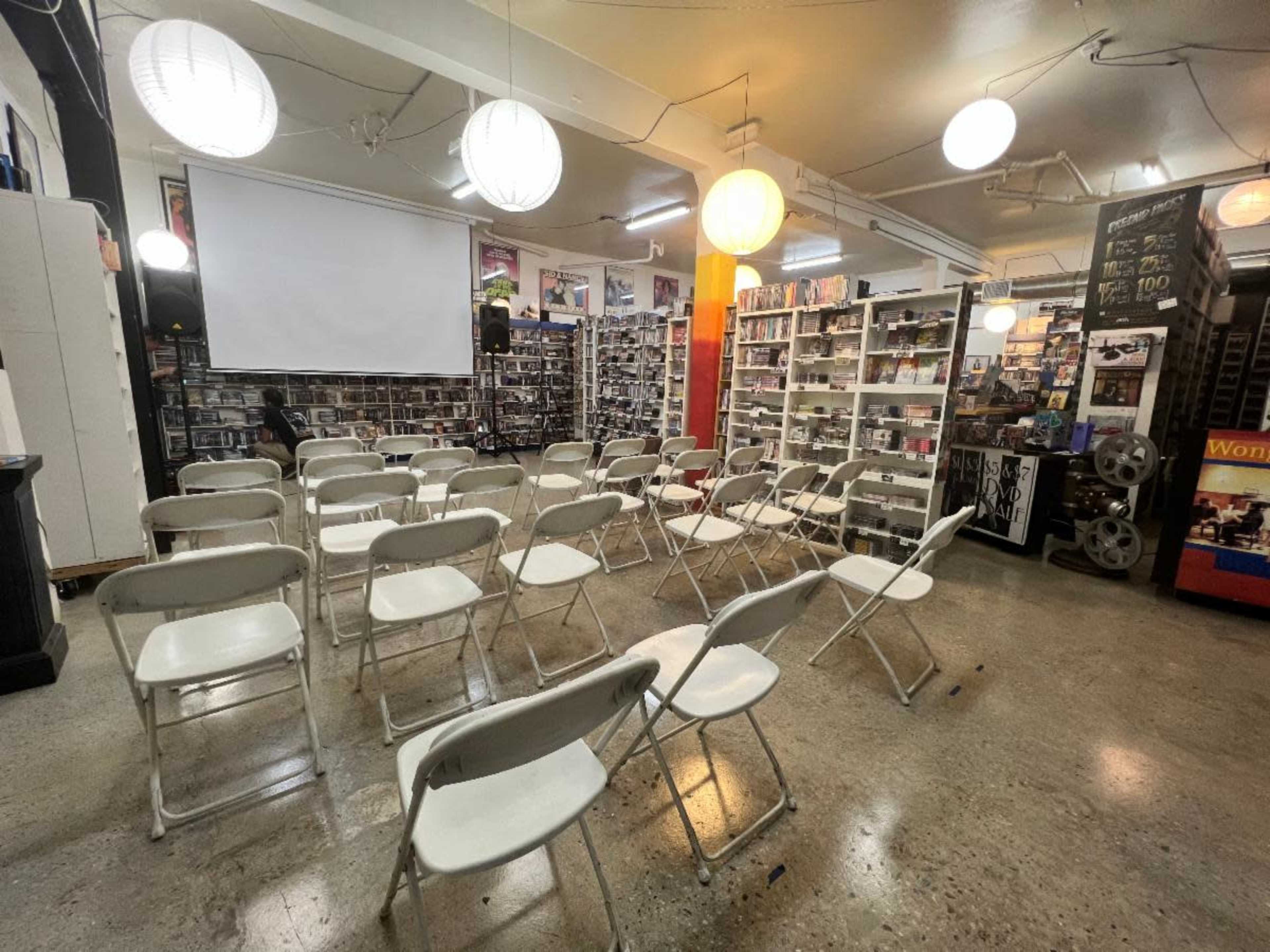 A room arranged with rows of white folding chairs facing a blank projector screen, surrounded by shelves filled with DVDs and other media.