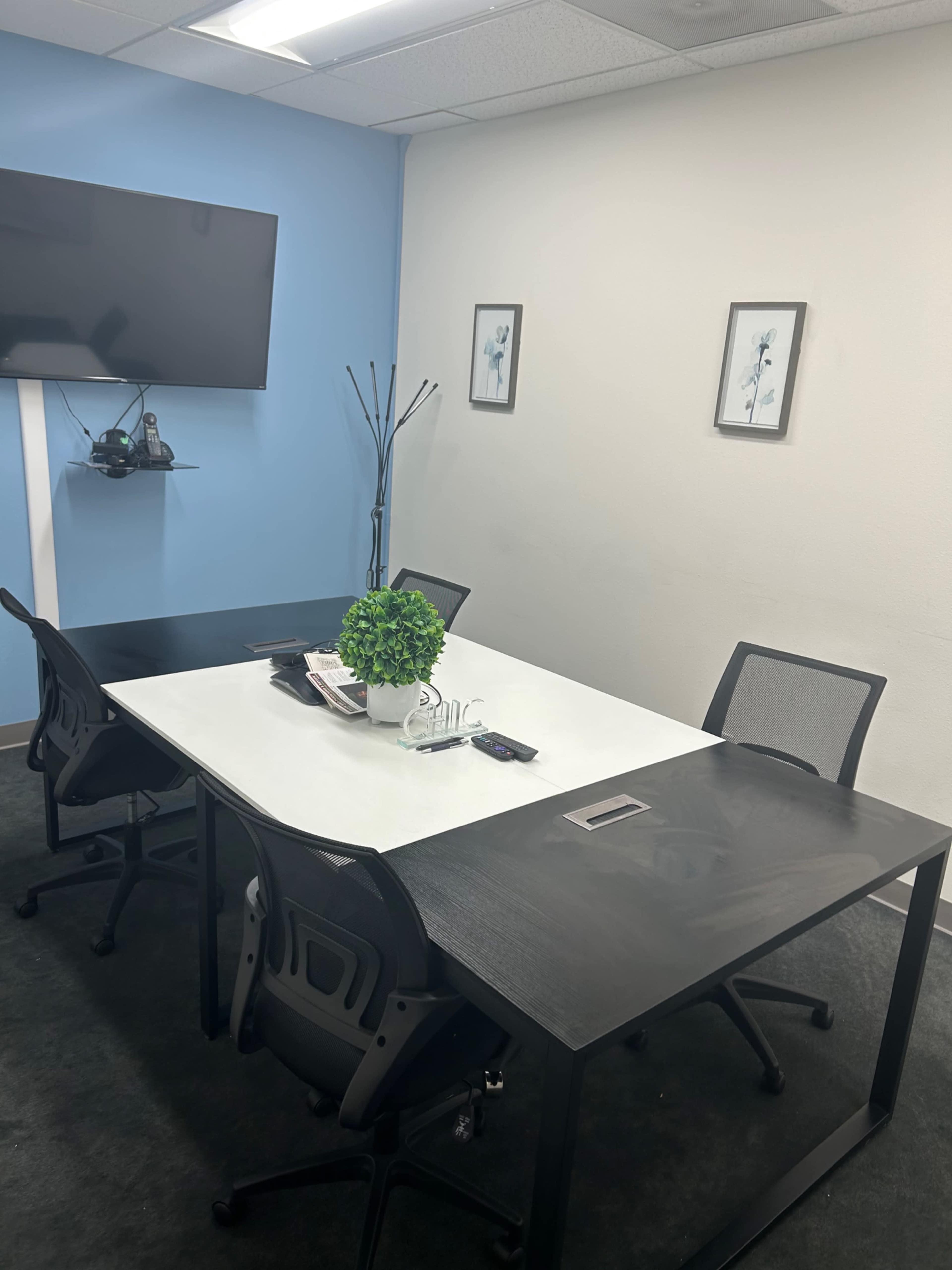 A small conference room with two rectangular tables, four chairs, a television on the wall, and two wall-mounted frames.