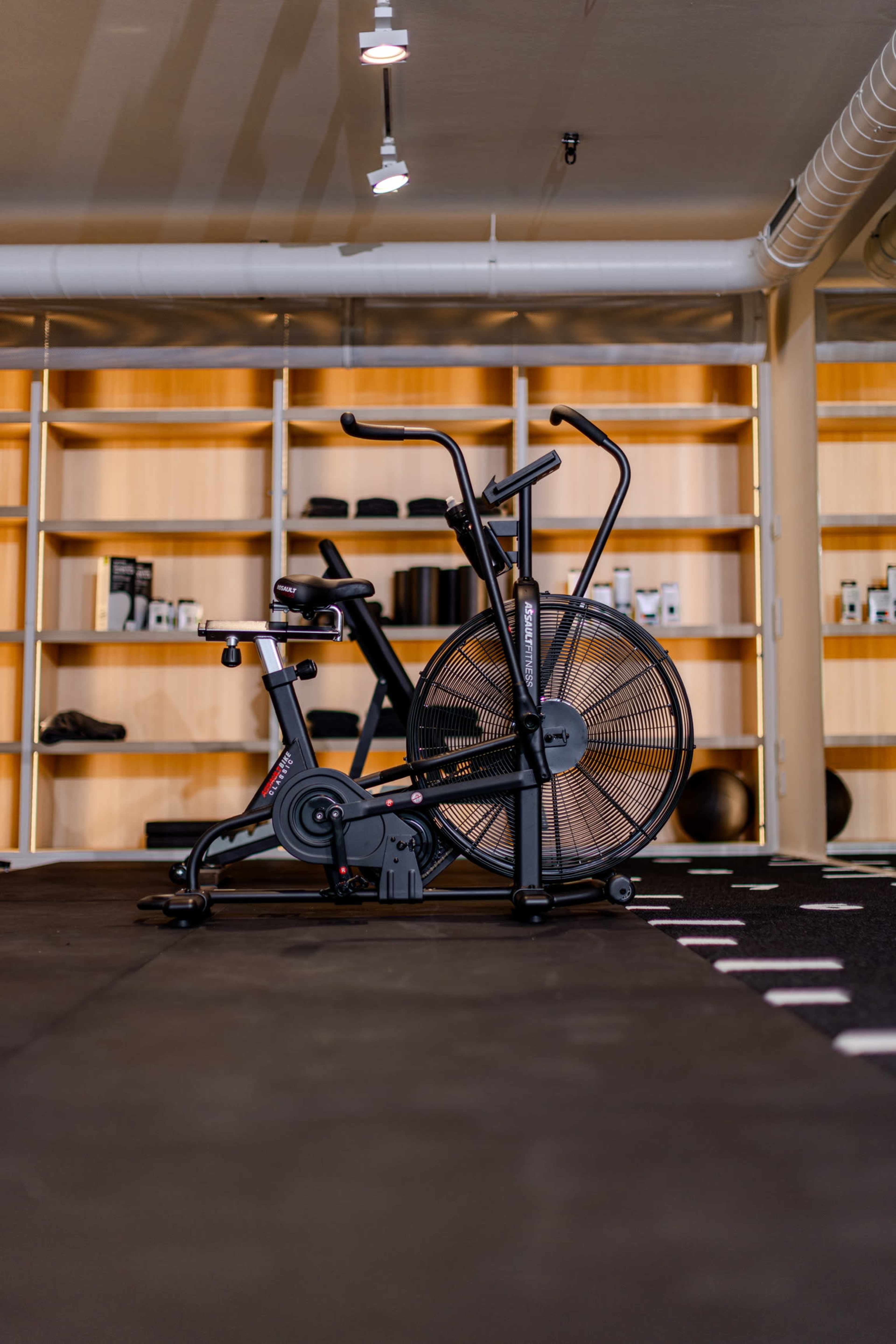The image shows a black exercise bike positioned on a mat in a gym with wooden shelves in the background holding various fitness products.