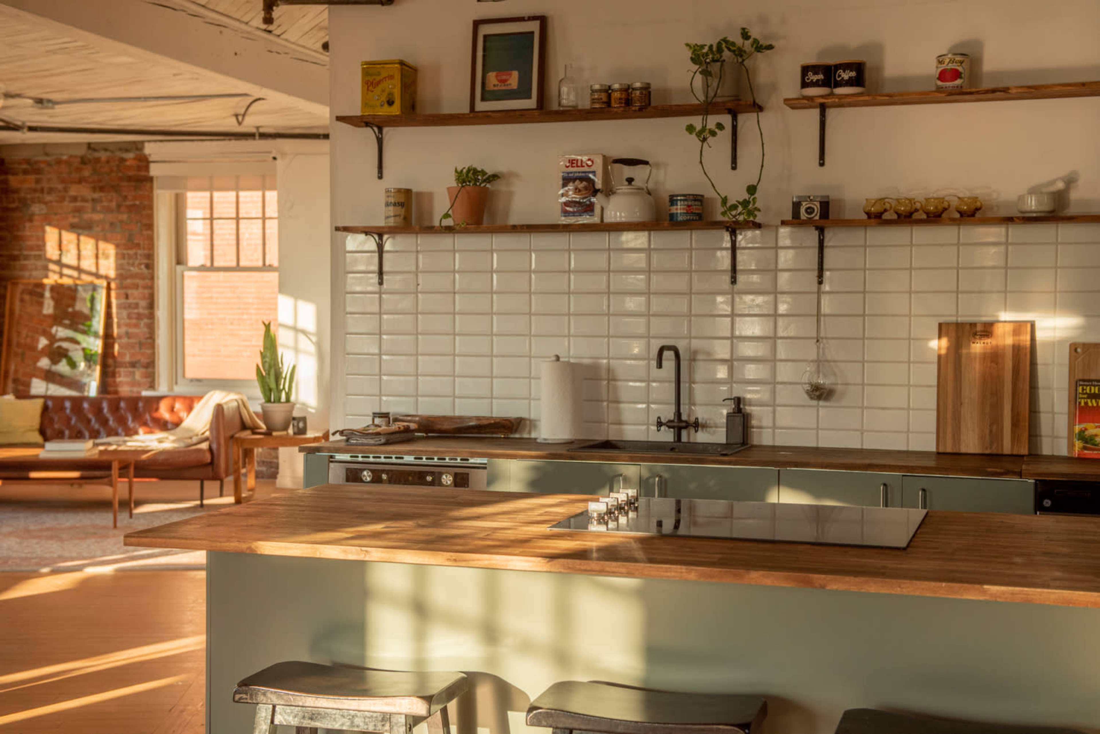 A bright and modern kitchen with wooden countertops, open shelving, and a large window casting light across the tiled wall.