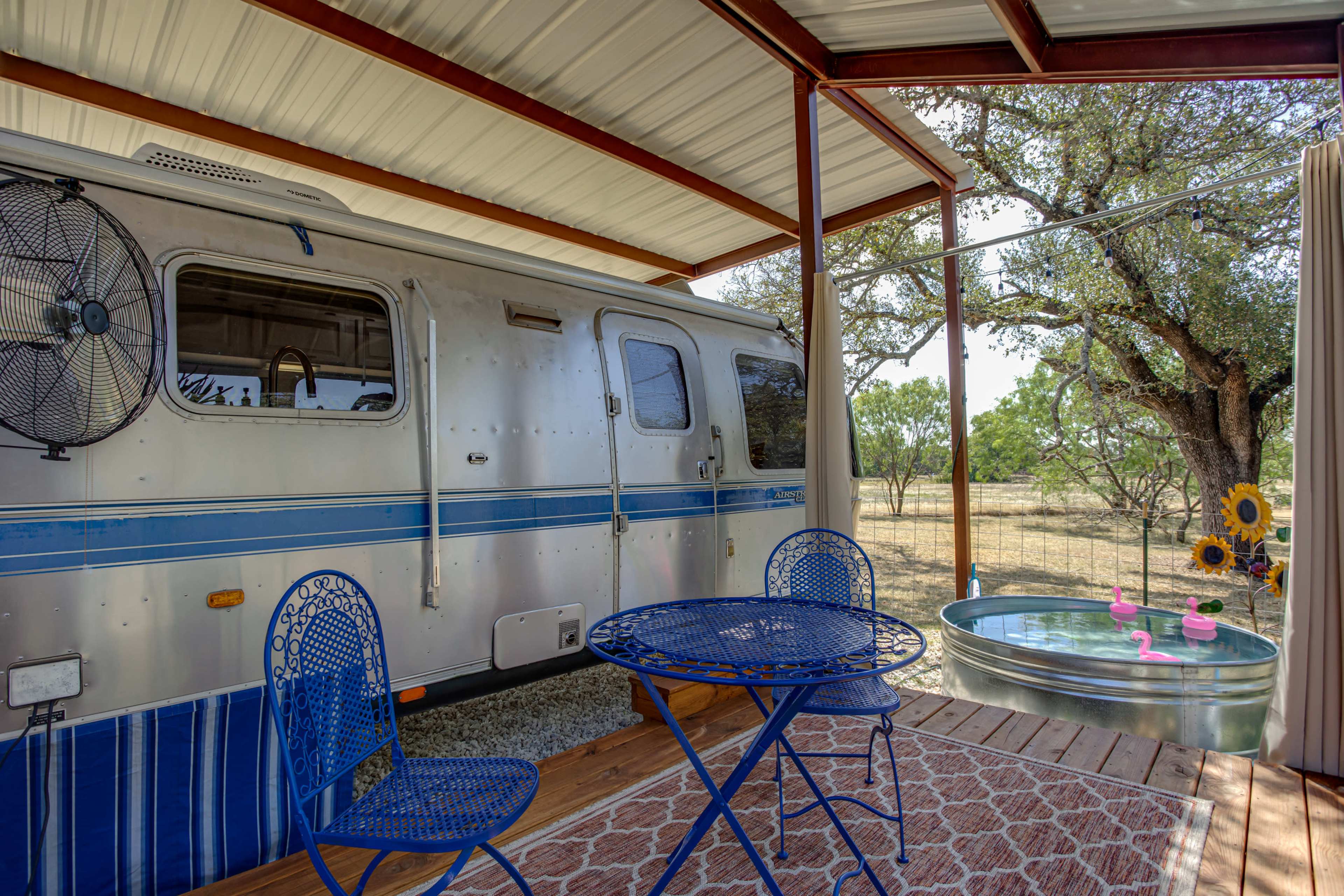 A blue and silver vintage trailer is parked under a metal awning next to a small round table with two chairs and a galvanized tub decorated with pink flamingo decor.